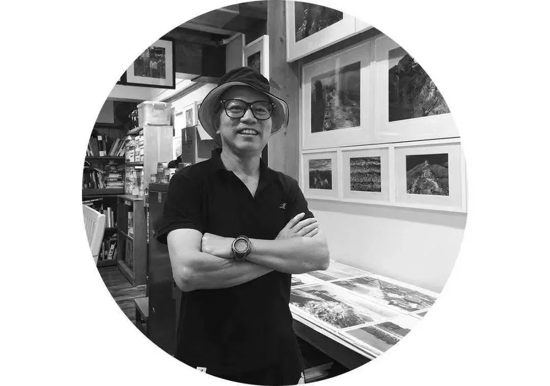 A man wearing glasses, a wide-brimmed hat, and a black polo shirt, standing with arms crossed in an art gallery or studio, surrounded by framed landscape photographs and art supplies.