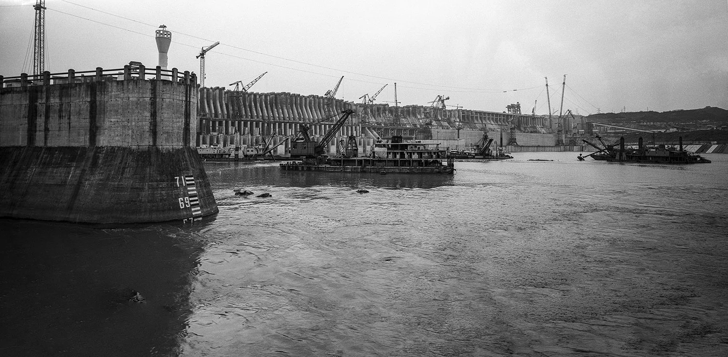 A black and white photo of a harbor with a large ship in the foreground, construction cranes, boats, and a dam structure in the background.