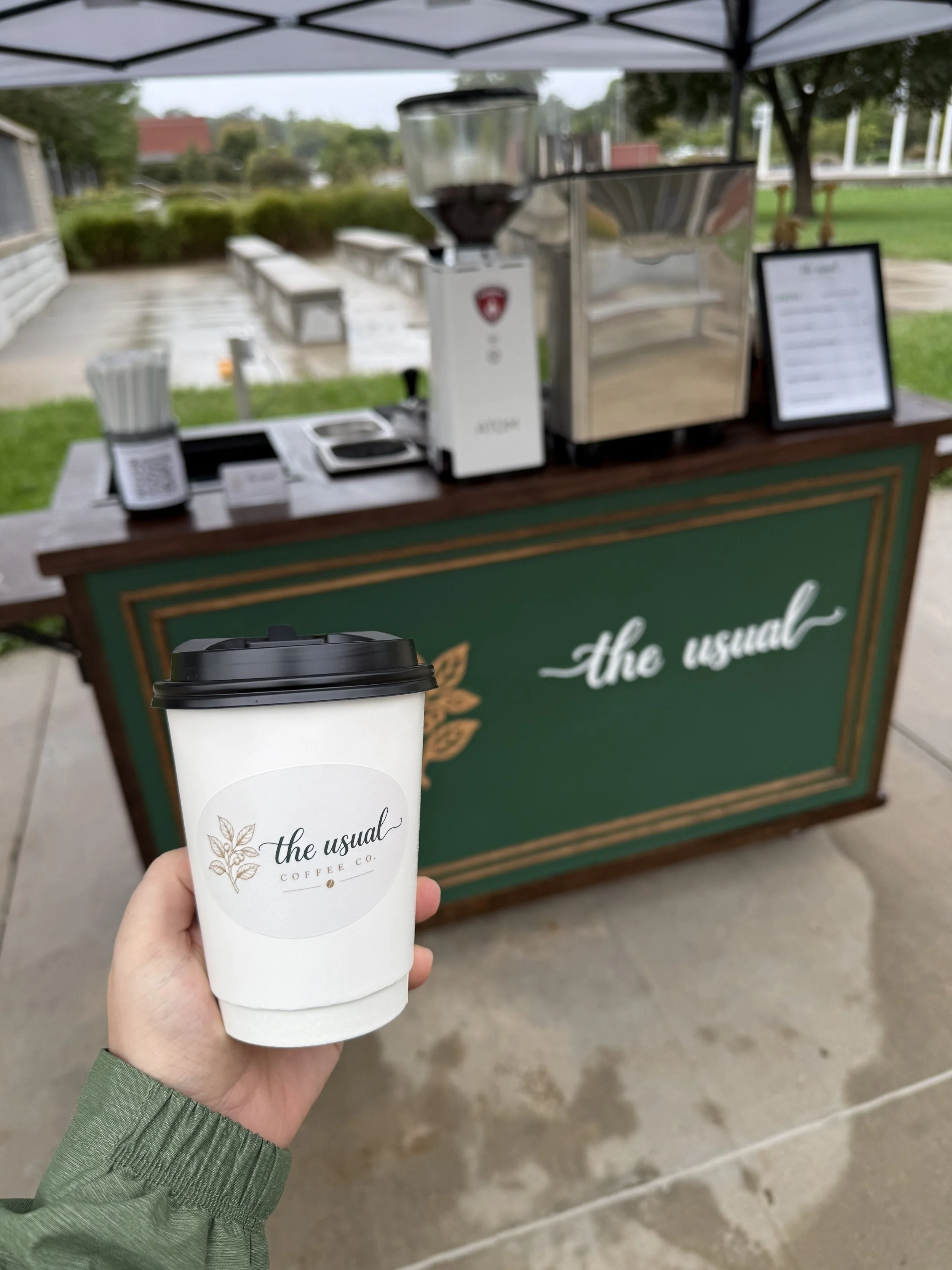 A branded hot coffee cup in front of The Usual Coffee Co. mobile espresso cart