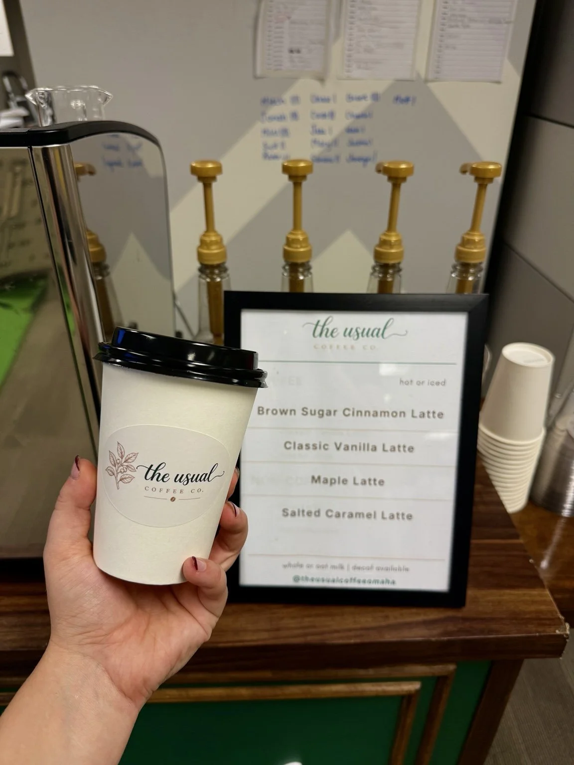 Branded hot coffee cup in front of The Usual Coffee Co. latte menu and syrup bottles.