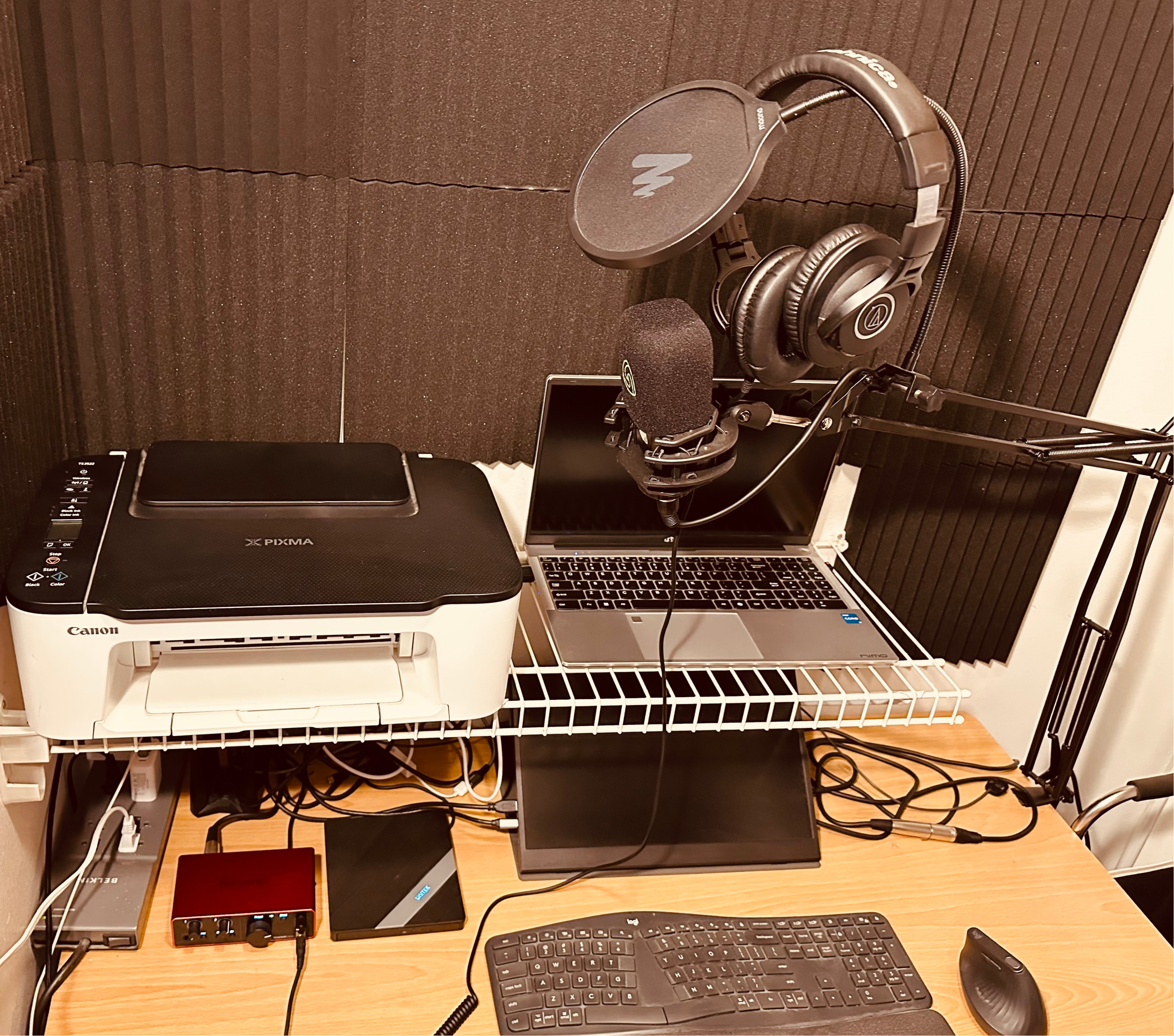A workspace with a black and silver microphone, a computer monitor, a keyboard, a mouse, a pair of headphones hanging on the microphone, a printer, and various electronic devices and cables on a wooden desk against a wall with acoustic foam panels.