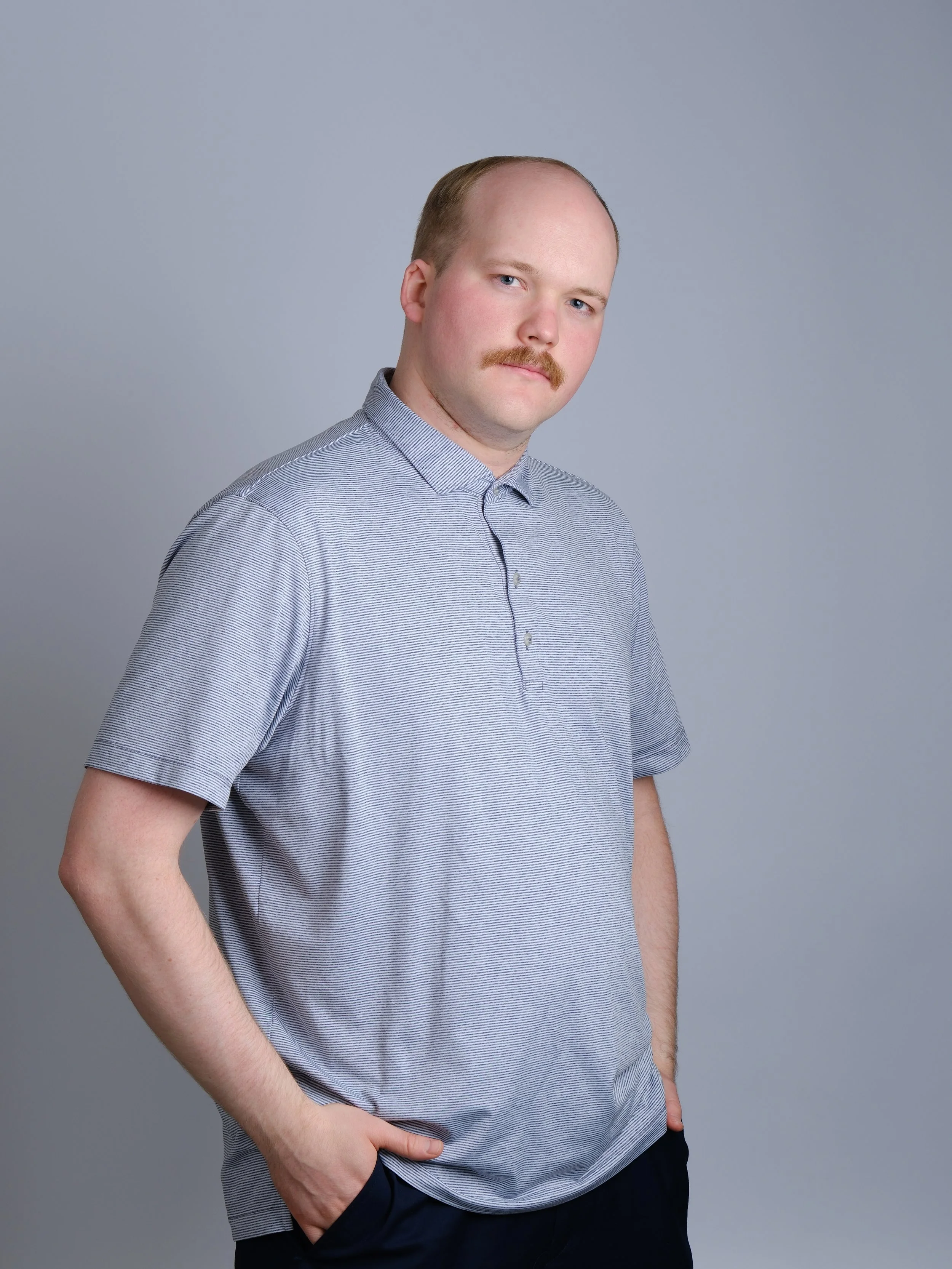 A young man with a mustache and light skin, wearing a short-sleeved, light-colored, striped shirt, standing against a plain gray background.