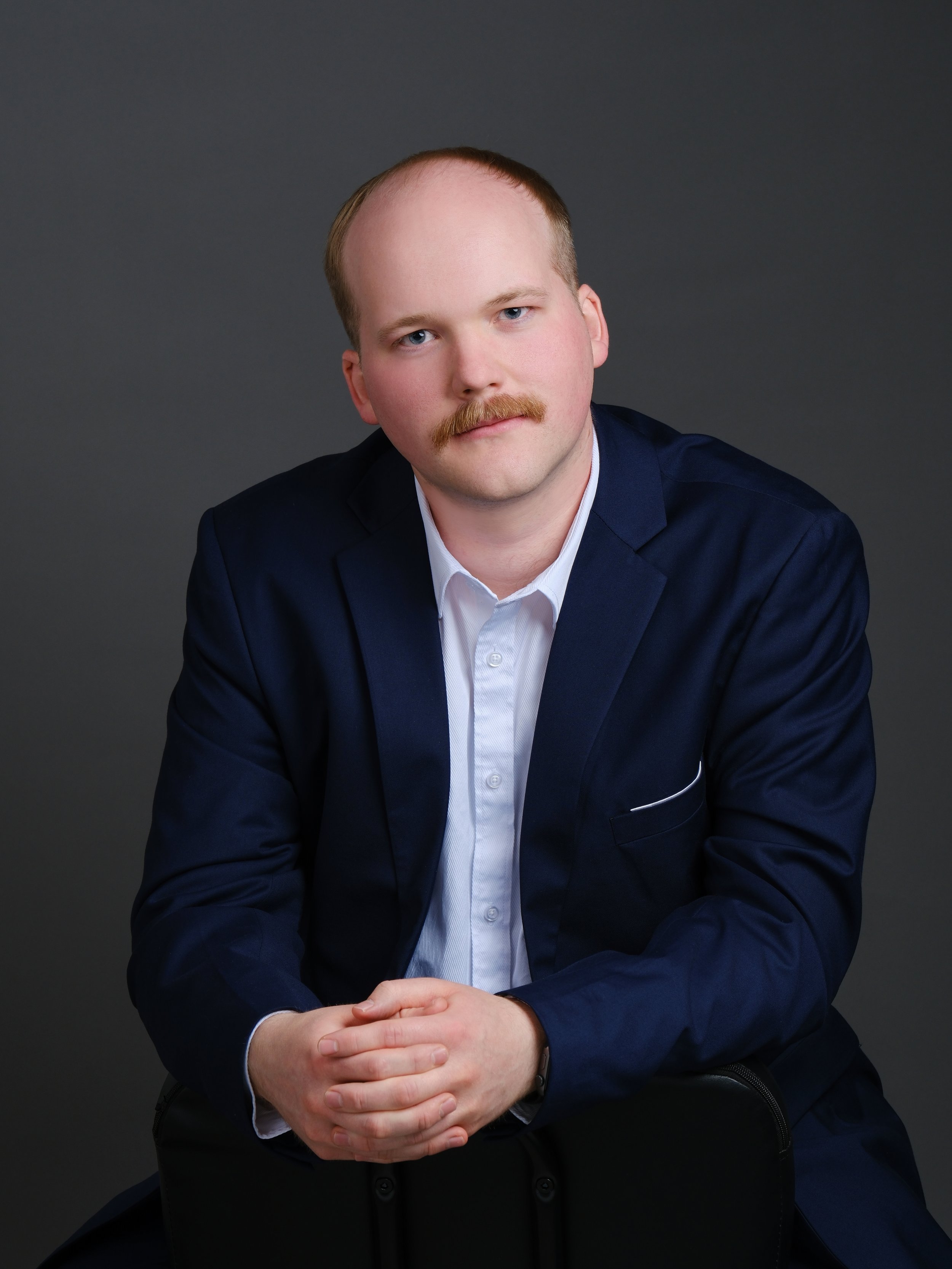 A man with a mustache wearing a dark blue blazer and white shirt, sitting with hands clasped, against a dark gray background.