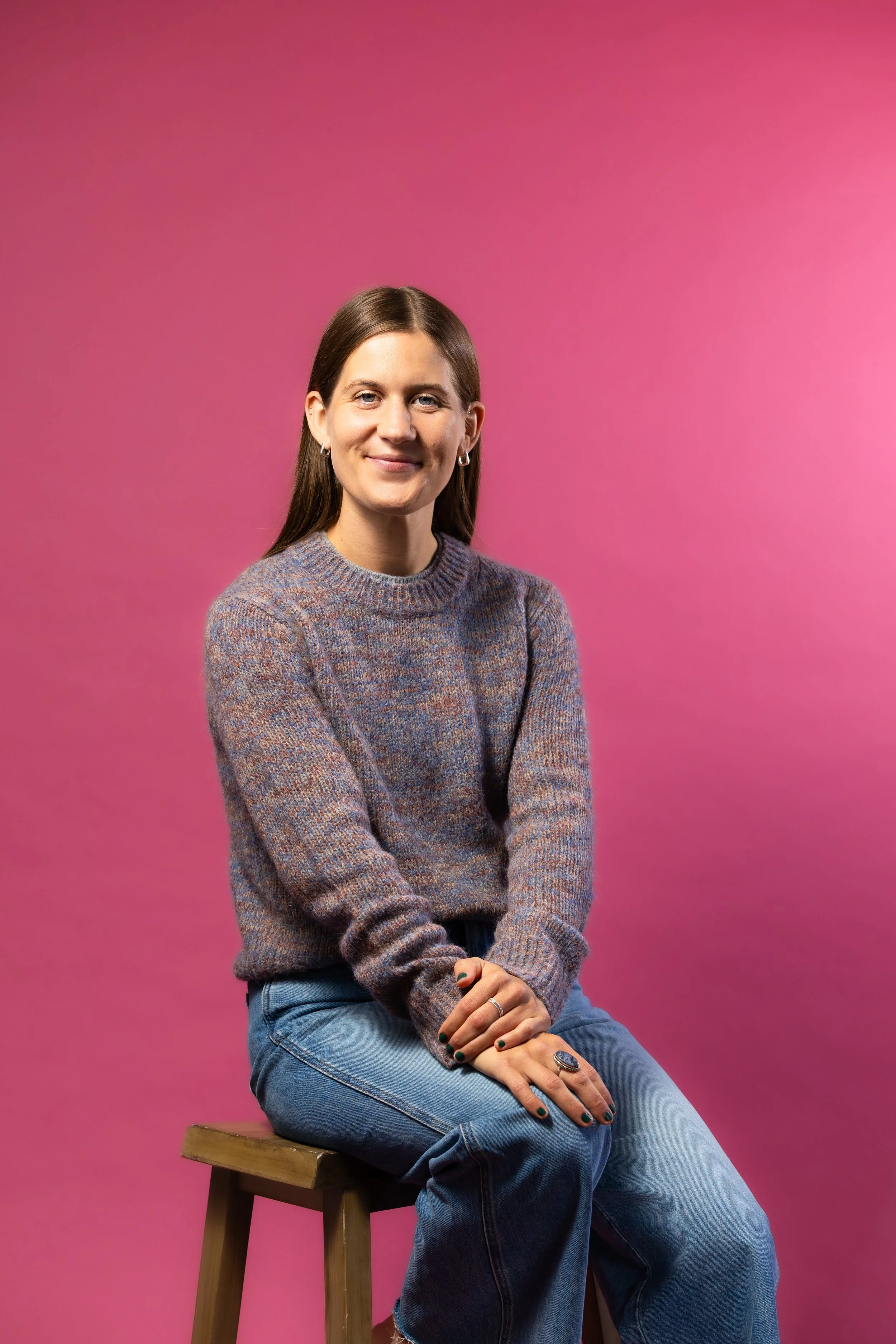 A woman sitting on a wooden stool against a pink background, wearing a gray sweater and blue jeans, with dark green nail polish and rings on her fingers, smiling softly.