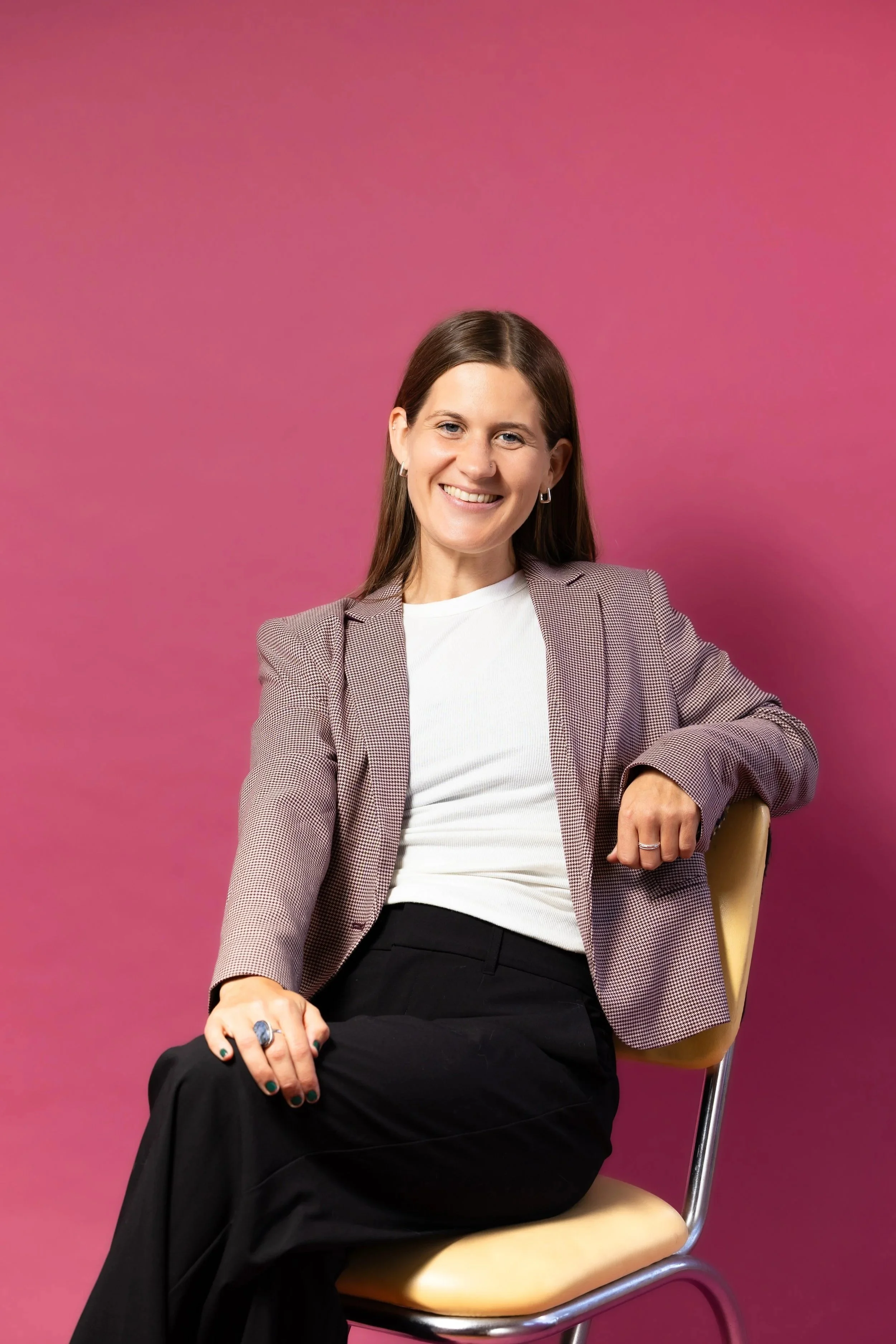 A smiling woman with brown hair, wearing a checked blazer, white t-shirt, black pants, and accessorized with earrings, rings, and dark nail polish, sitting on a beige chair against a solid pink background.