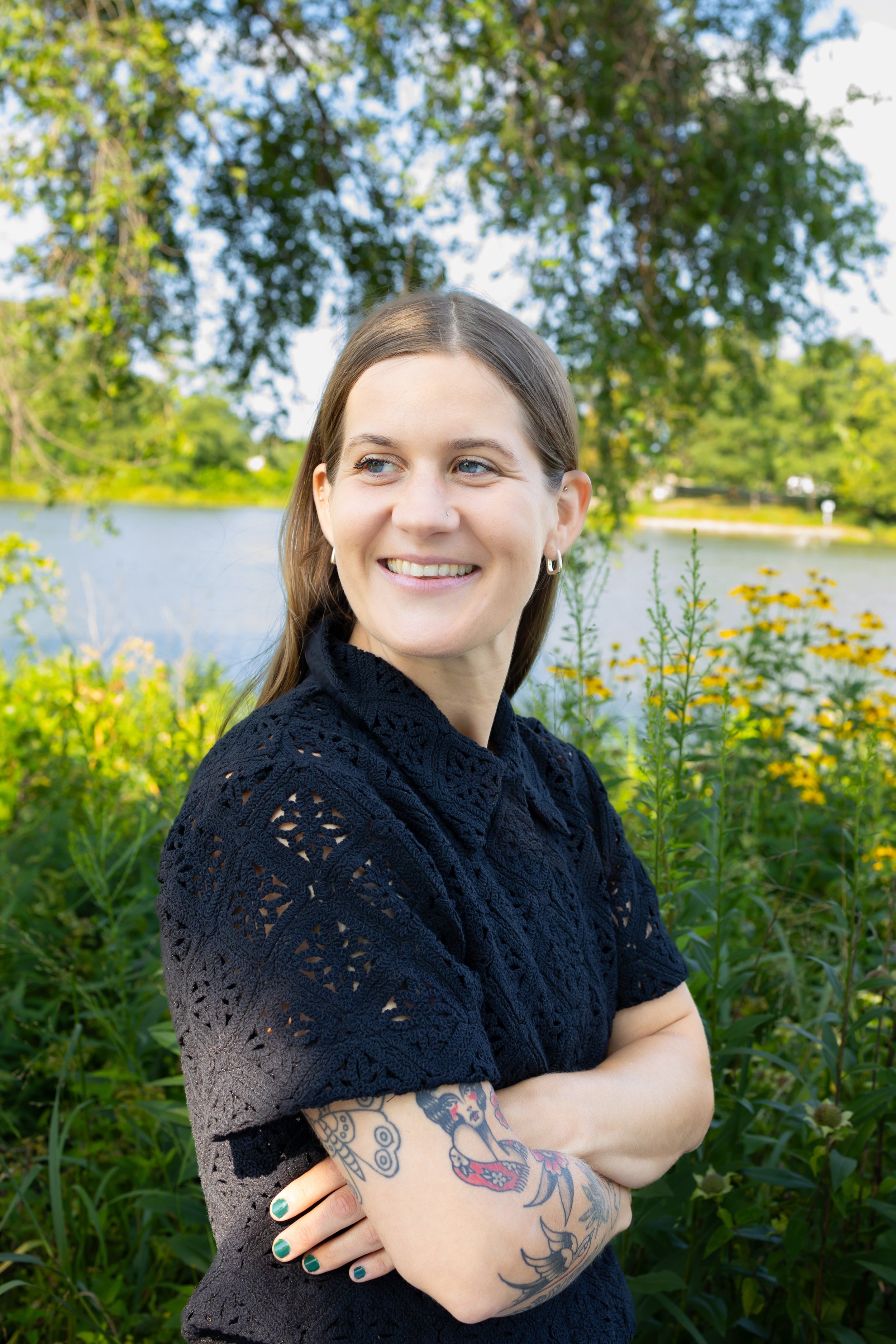 A young woman with long brown hair and tattoos on her arm, smiling and standing with arms crossed in front of a river with trees and plants in the background.