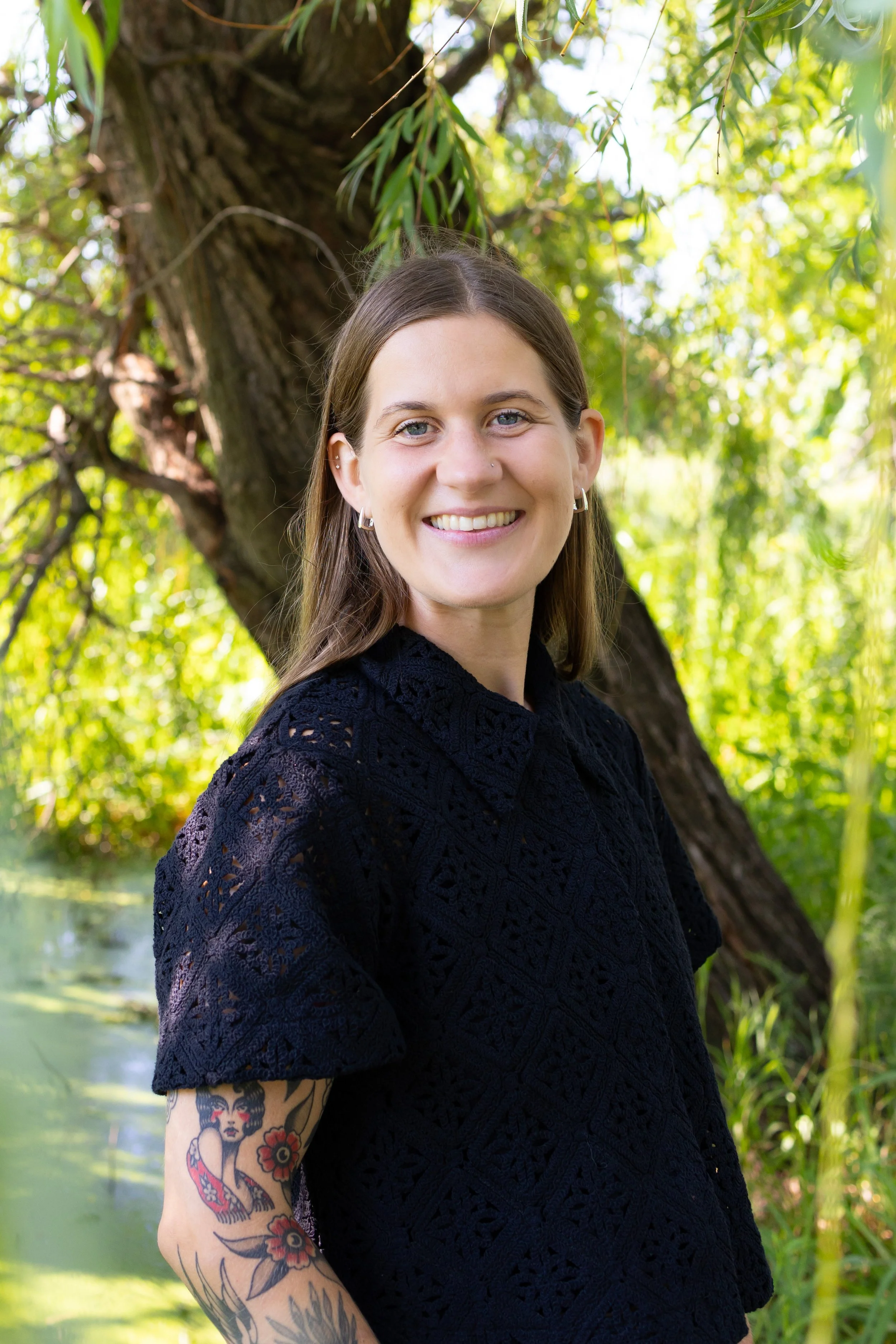 A smiling woman with blue eyes, dark brown hair, and multiple earrings standing outdoors near a large tree with green leaves and water in the background.