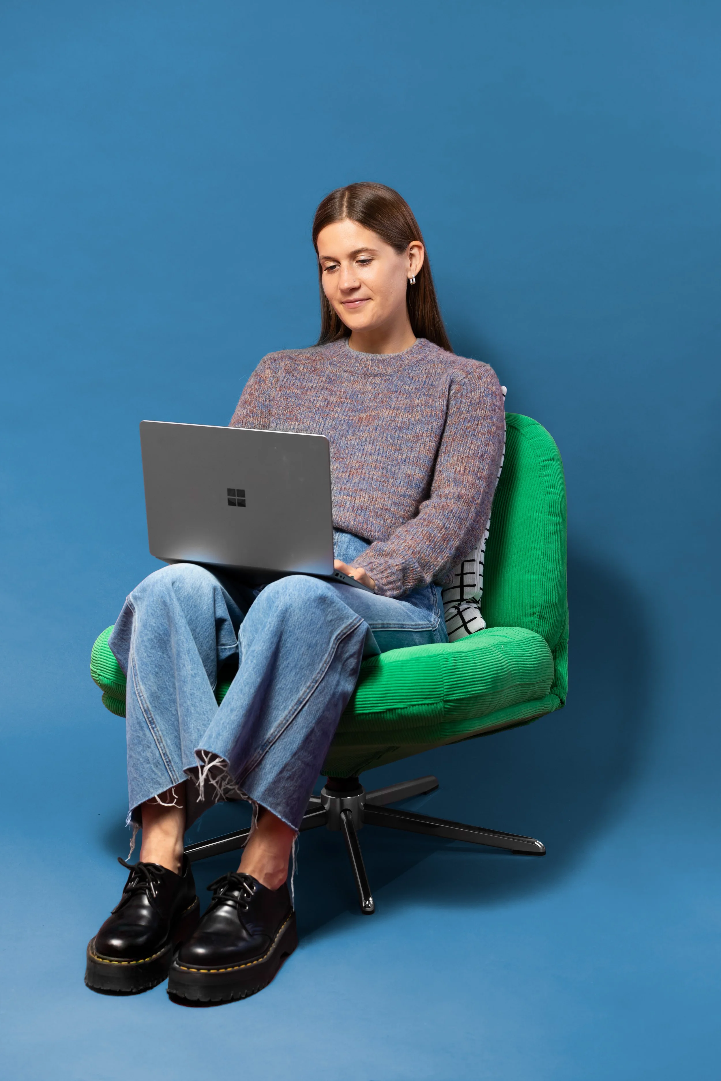 A young woman sitting on a green office chair against a blue background, looking at a laptop resting on her lap, wearing a multicolored sweater, light blue jeans, black shoes, and earrings.