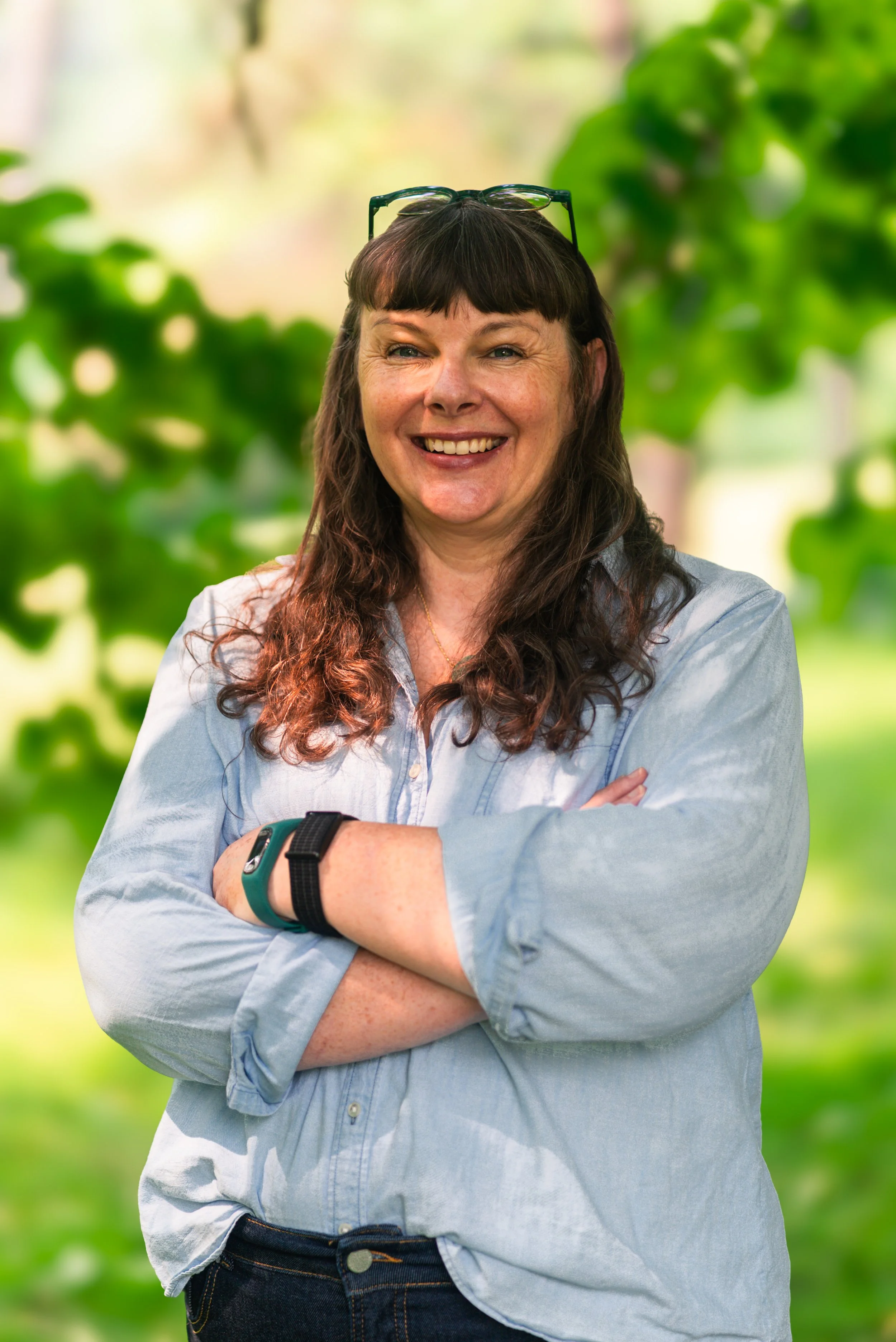 A woman with long brown wavy hair and bangs, smiling and crossing her arms, wearing a light blue button-up shirt and black jeans, standing outdoors in front of blurred green foliage.