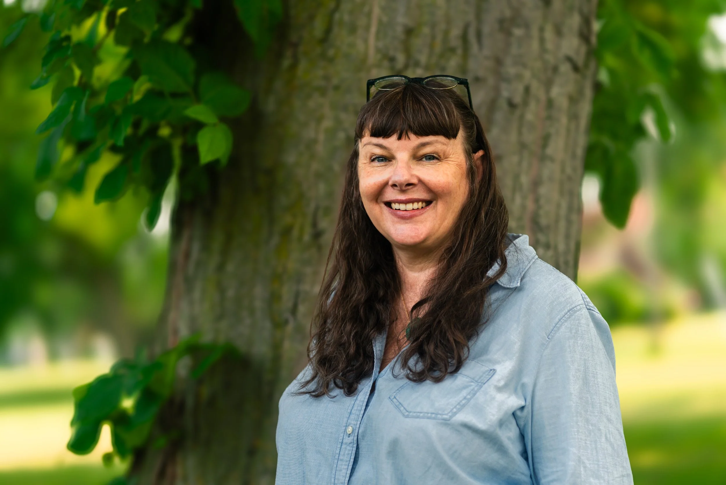 A smiling woman with long brown hair and glasses on her head, wearing a light blue denim shirt, standing outdoors near a tree with green leaves.