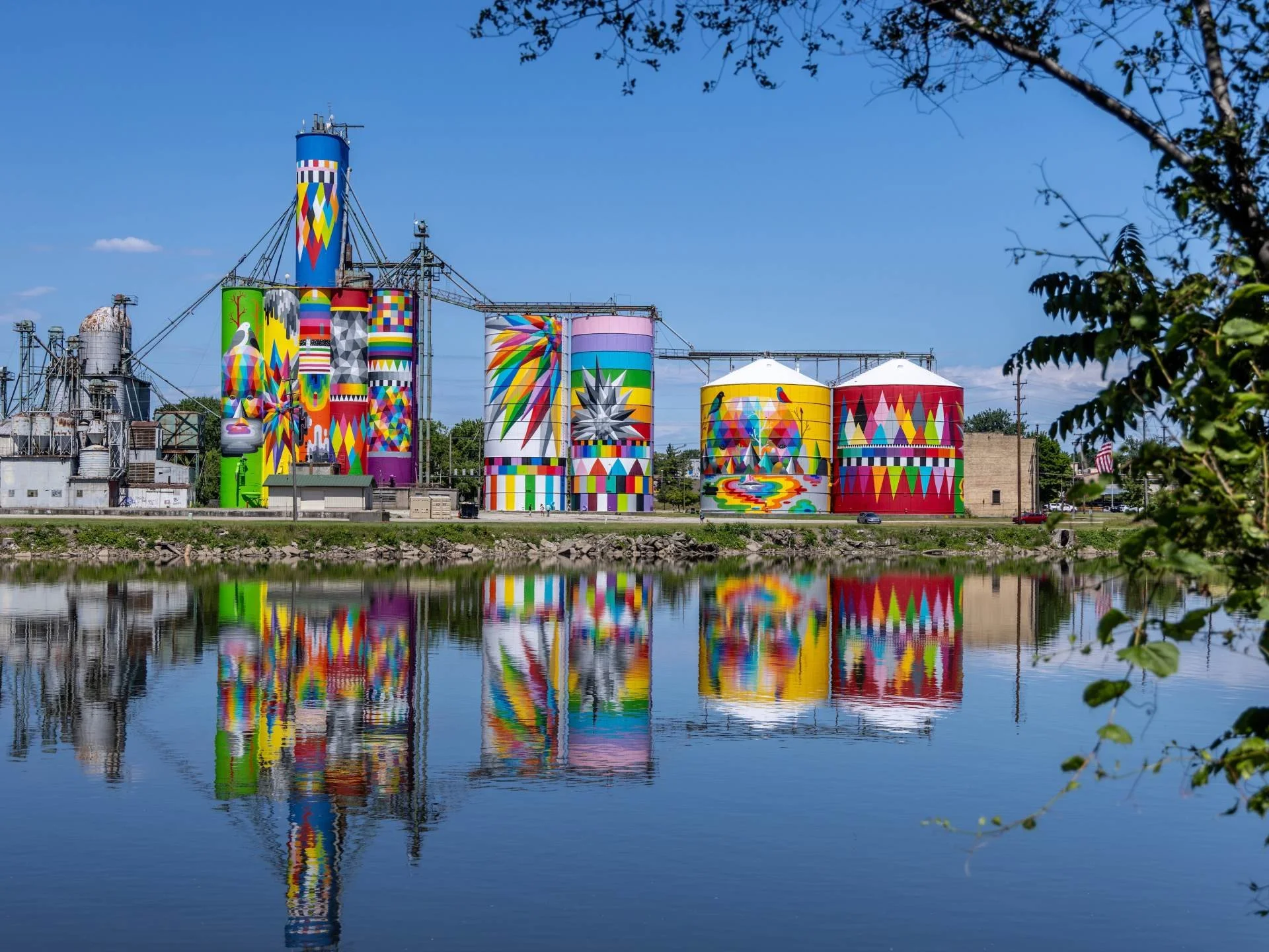Colorful industrial silos with rainbow and geometric patterns, reflected on a calm river, with trees and blue sky in the background.