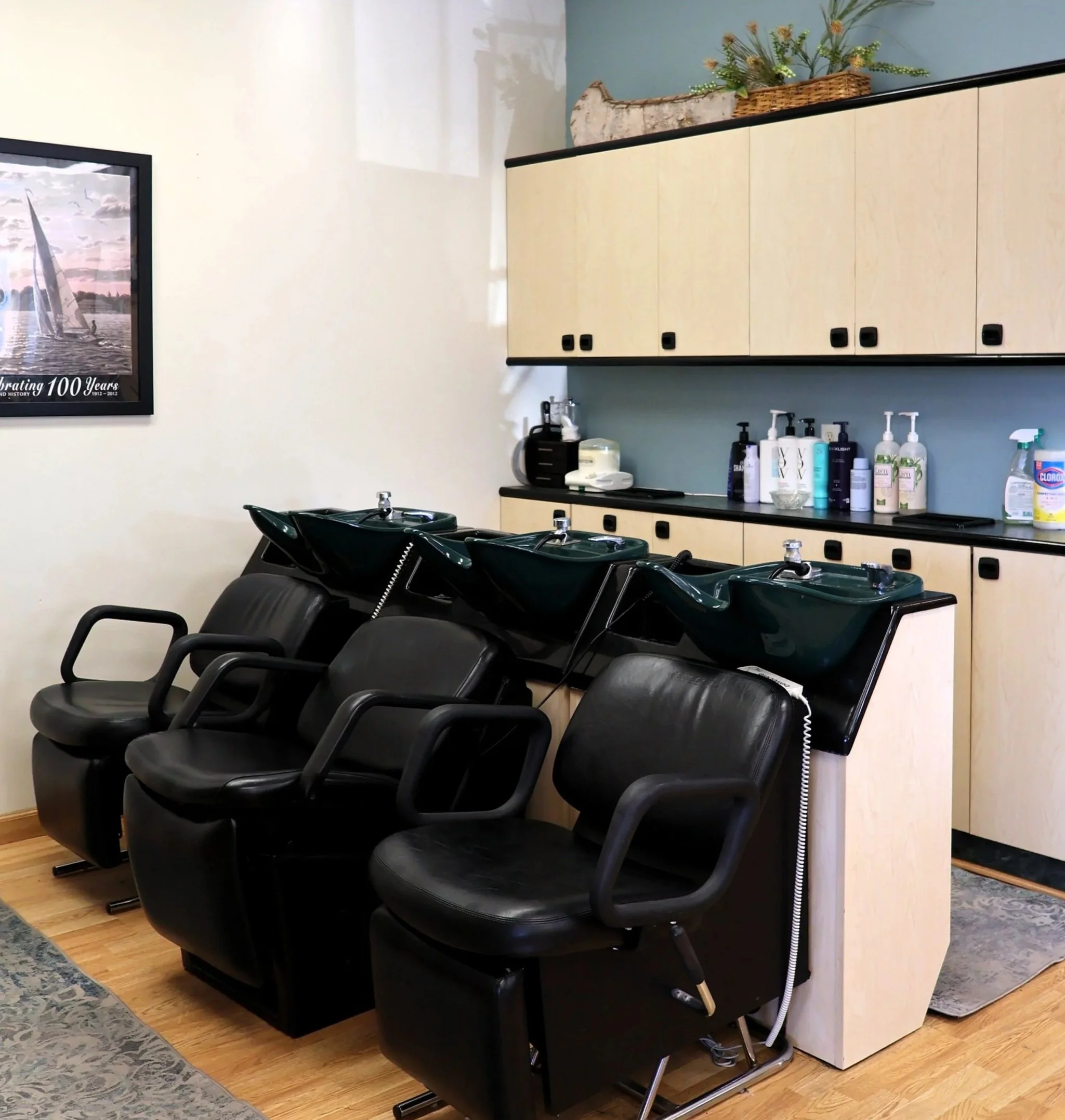 Salon or spa room with black styling chairs in front of a mirror and a light-colored cabinet with soap and lotion bottles on the counter.