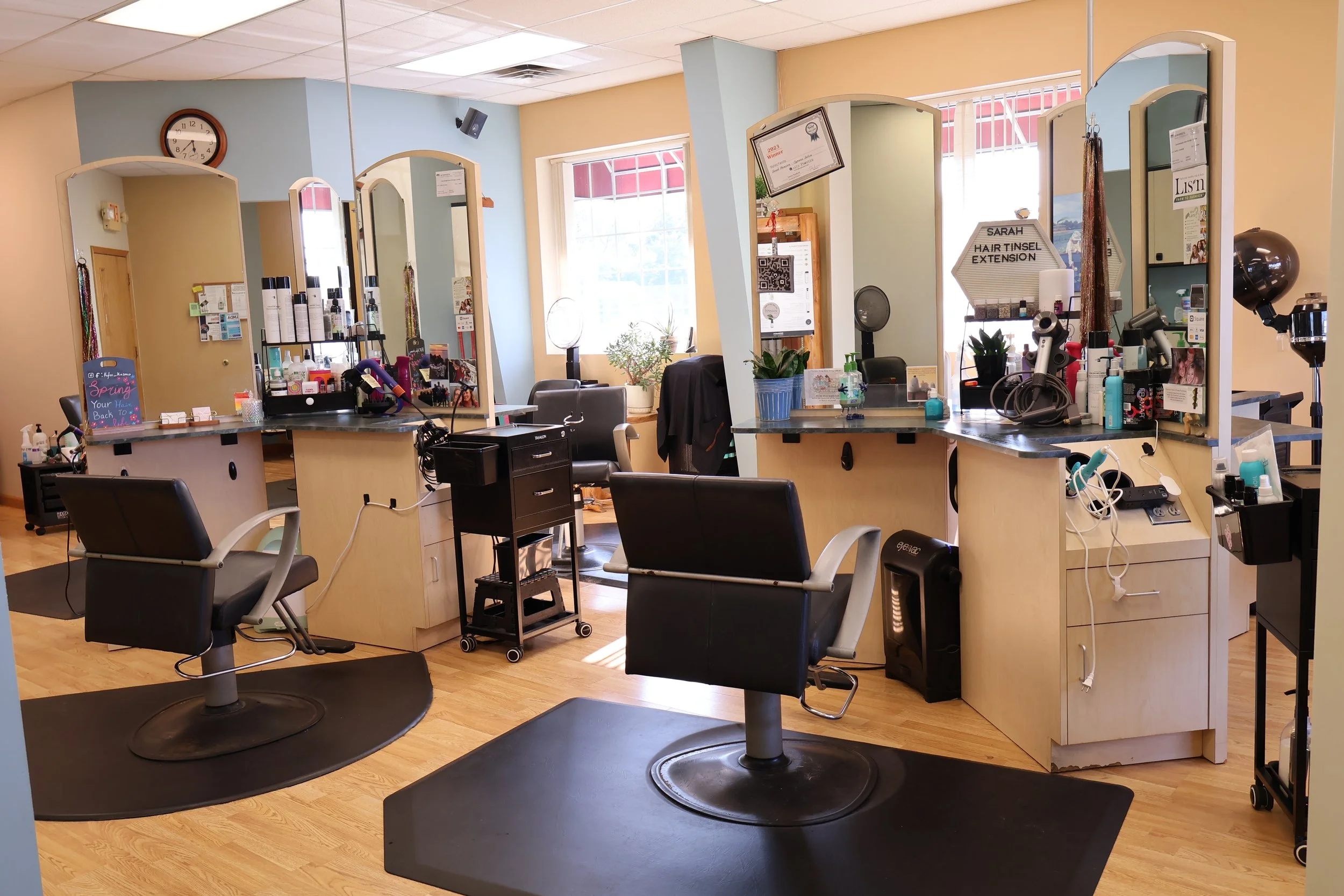 Hairstyling salon with multiple mirrors, chairs, and hair styling equipment. Natural light from large windows, wooden flooring, and various hair care products on countertops.