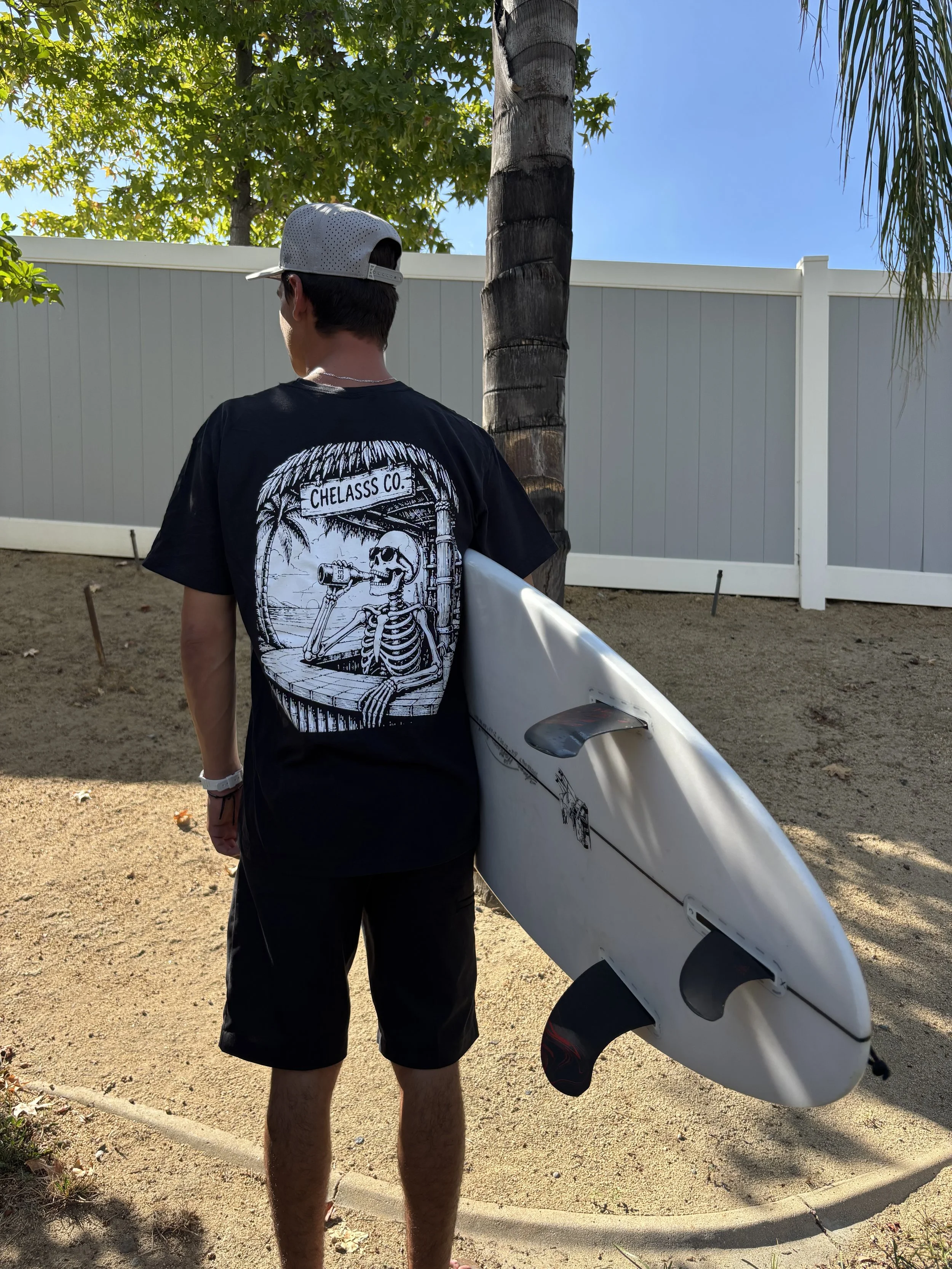 Back view of a young man wearing a white baseball cap, black t-shirt with a skeleton design, and black shorts, holding a white surfboard, standing on a sandy area near a tall palm tree and green foliage, with a white fence and blue sky in the background.