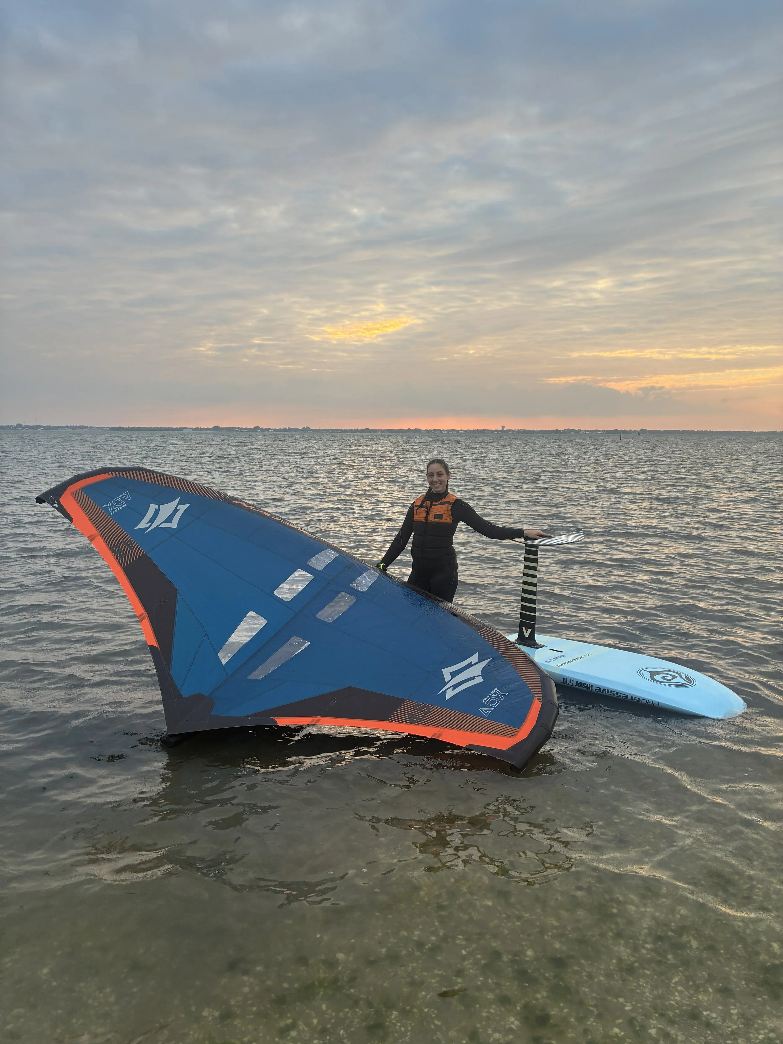 A woman, Katie from Katie's Kitewear, standing in shallow water at sunset, holding a wing for wingfoiling and a foil board in the water next to her. Related to kiteboarding and watersports