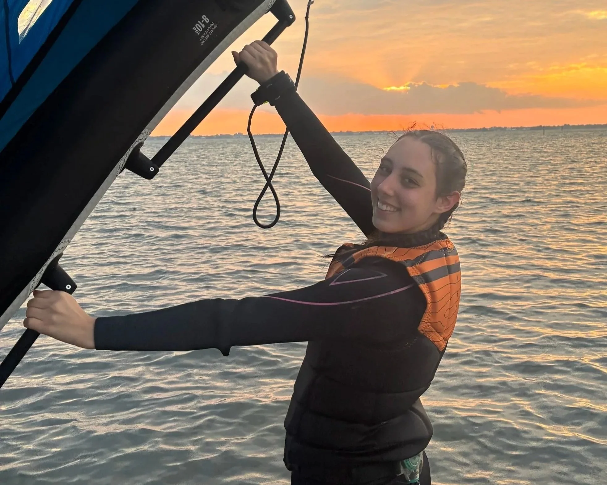 A young woman, Katie from Katie's Kitewear, in a wetsuit holds onto a wing for wingfoiling in the water during sunset, smiling at the camera. Related to kiteboarding