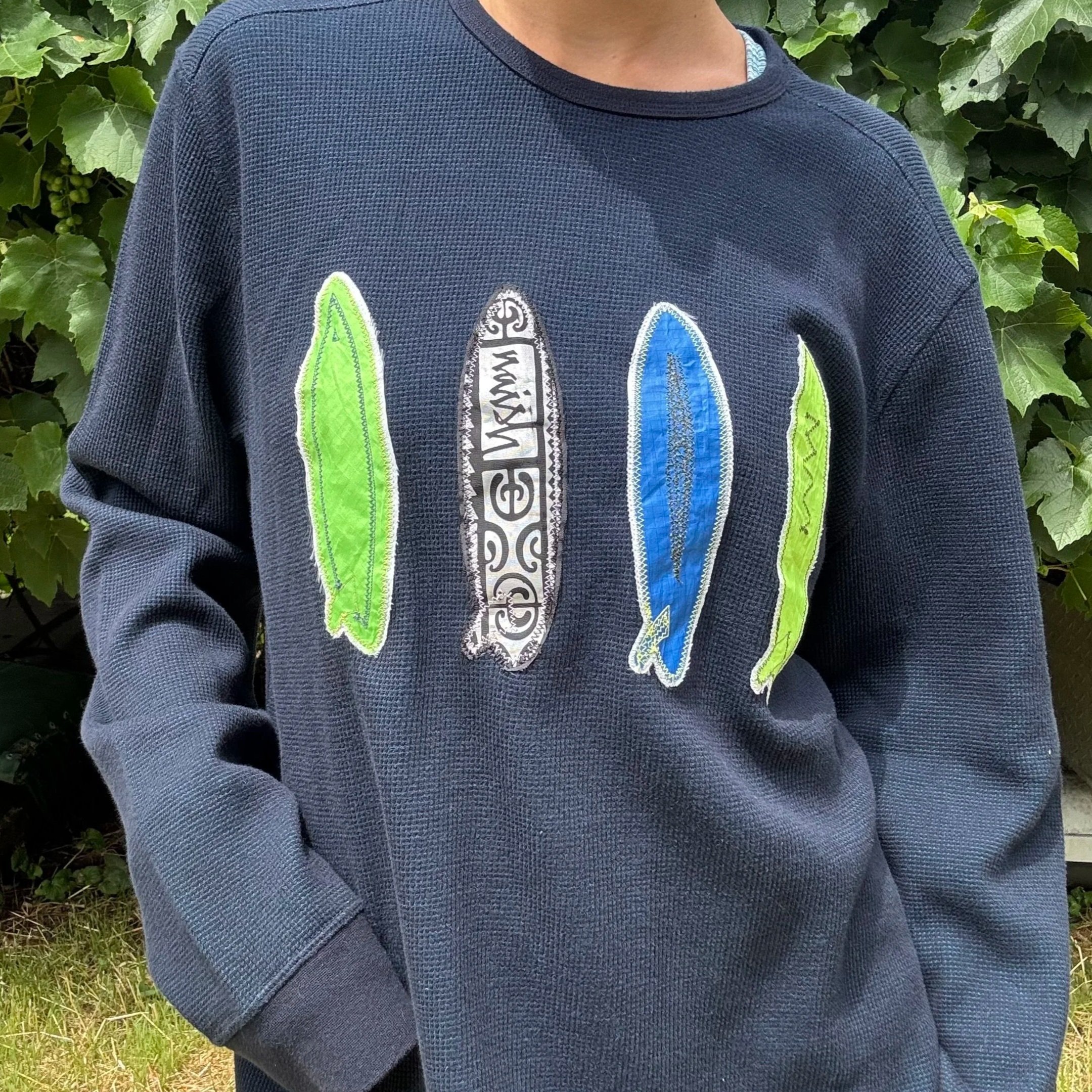 Close-up of a person wearing a navy blue sweatshirt with colorful embroidered surfboard designs, standing outdoors with green leafy plants in the background.