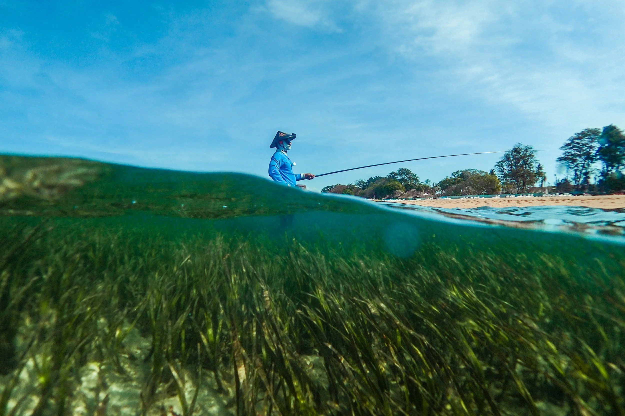 View of a person in blue standing on the shore holding a fishing rod, with a partly submerged camera, taken from underwater, showing water and grass underwater and trees and beach on land in the background.