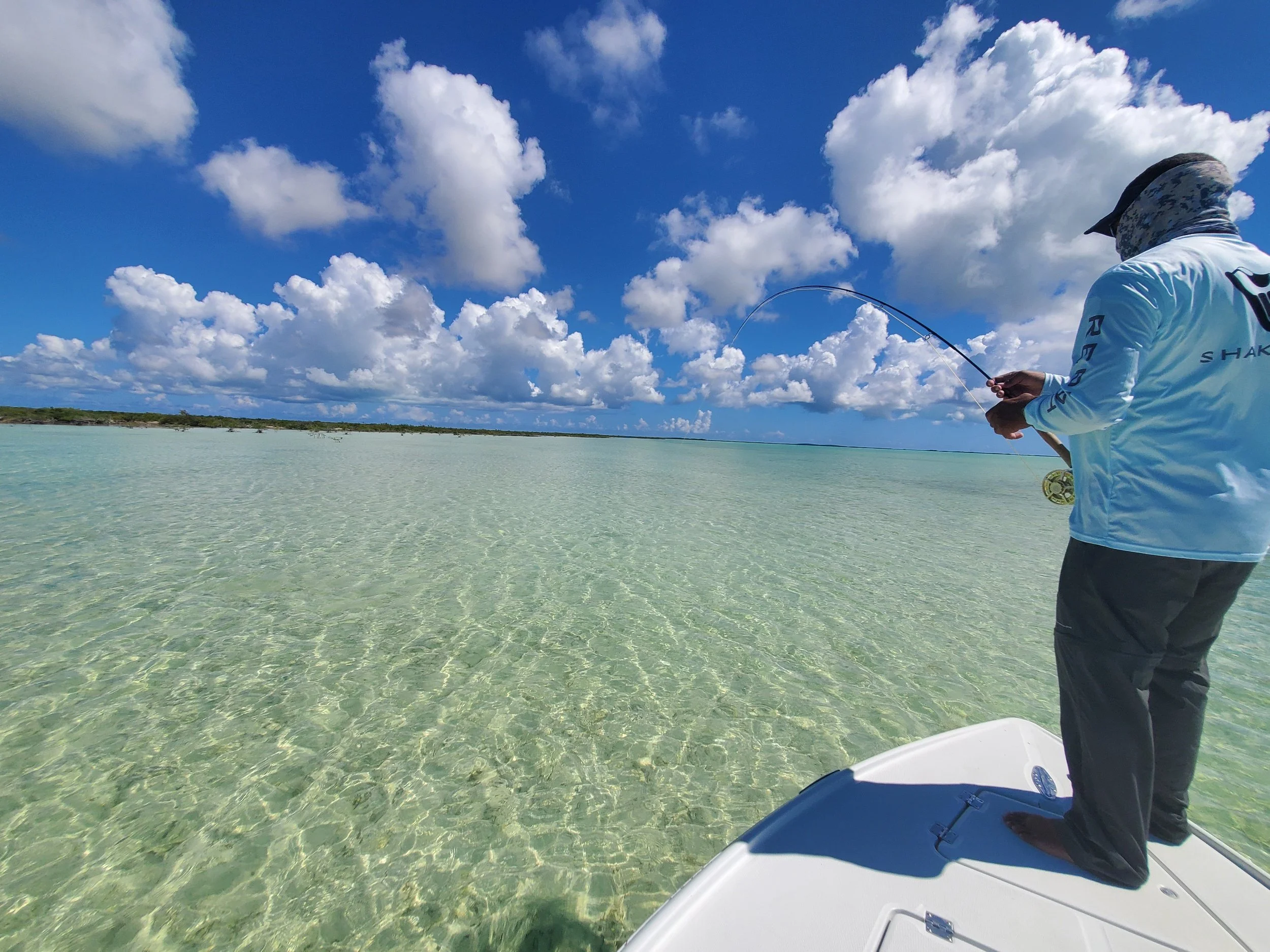 A person wearing a white shirt and a hat fishing from a boat in shallow, clear water under a blue sky with scattered clouds.