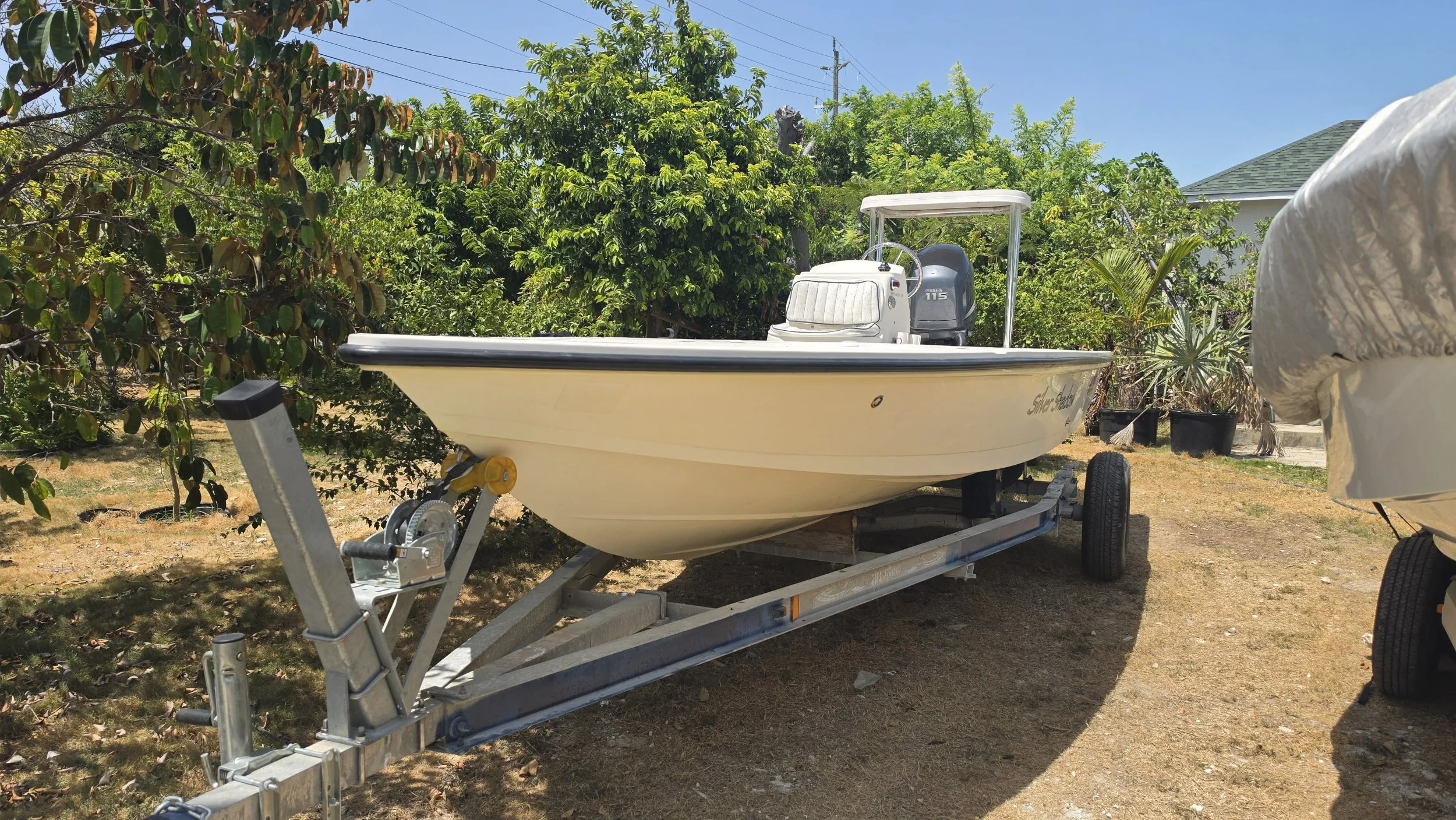 A small white boat on a trailer parked on dry grass, surrounded by lush green trees and plants under a clear blue sky.