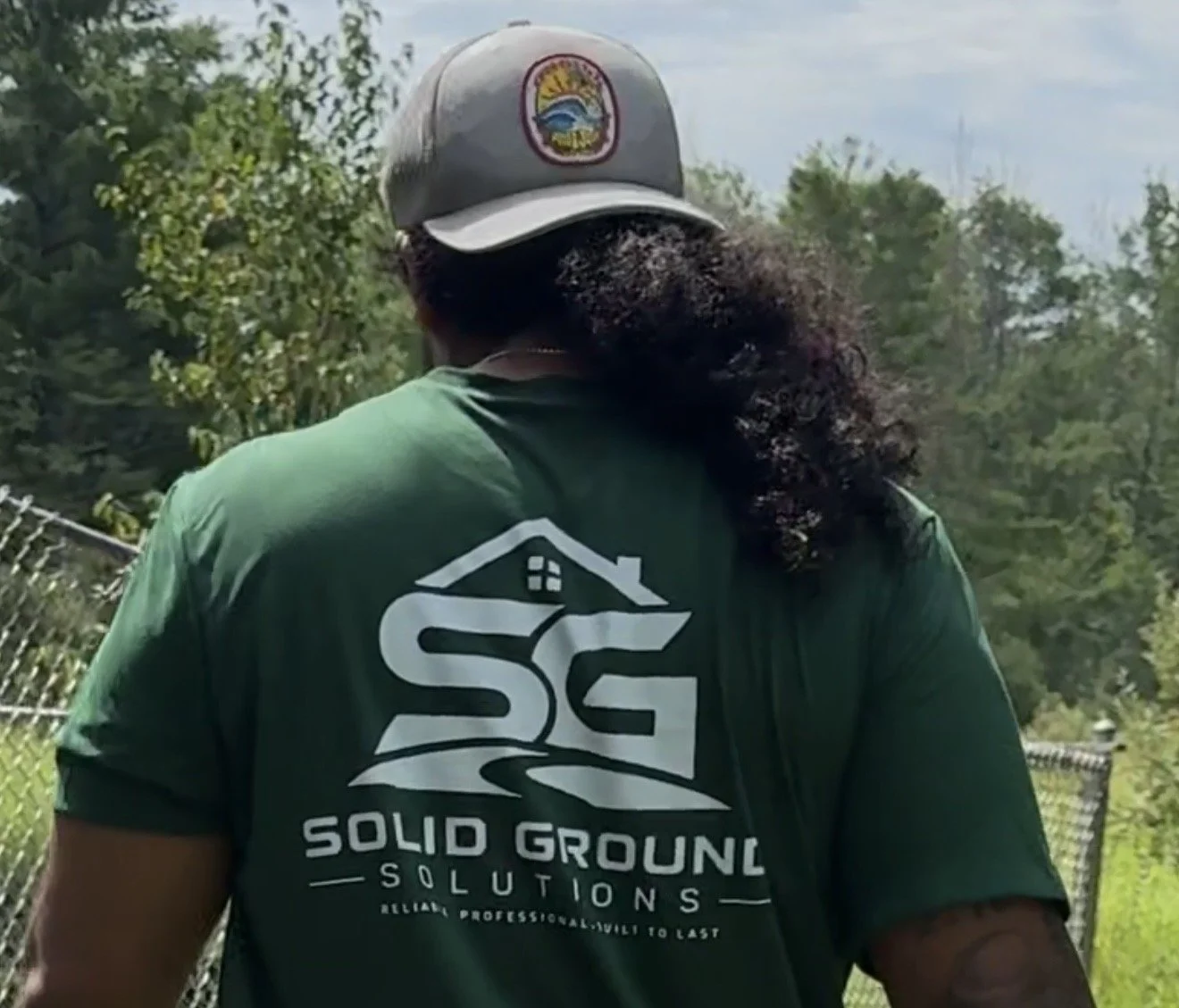 Person seen from back wearing a green t-shirt with the logo and text 'Solid Ground Solutions' and a gray cap with a logo, standing outdoors near a fence with trees in the background.