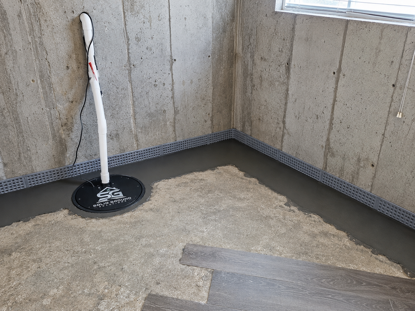 An unfinished room with concrete floors and walls, with a floor drain installed in the corner. There is a metal cove base around the perimeter, and a section of flooring has been freshly poured with gray cement. An electric pipe or pole with a charging station label is plugged into the wall.