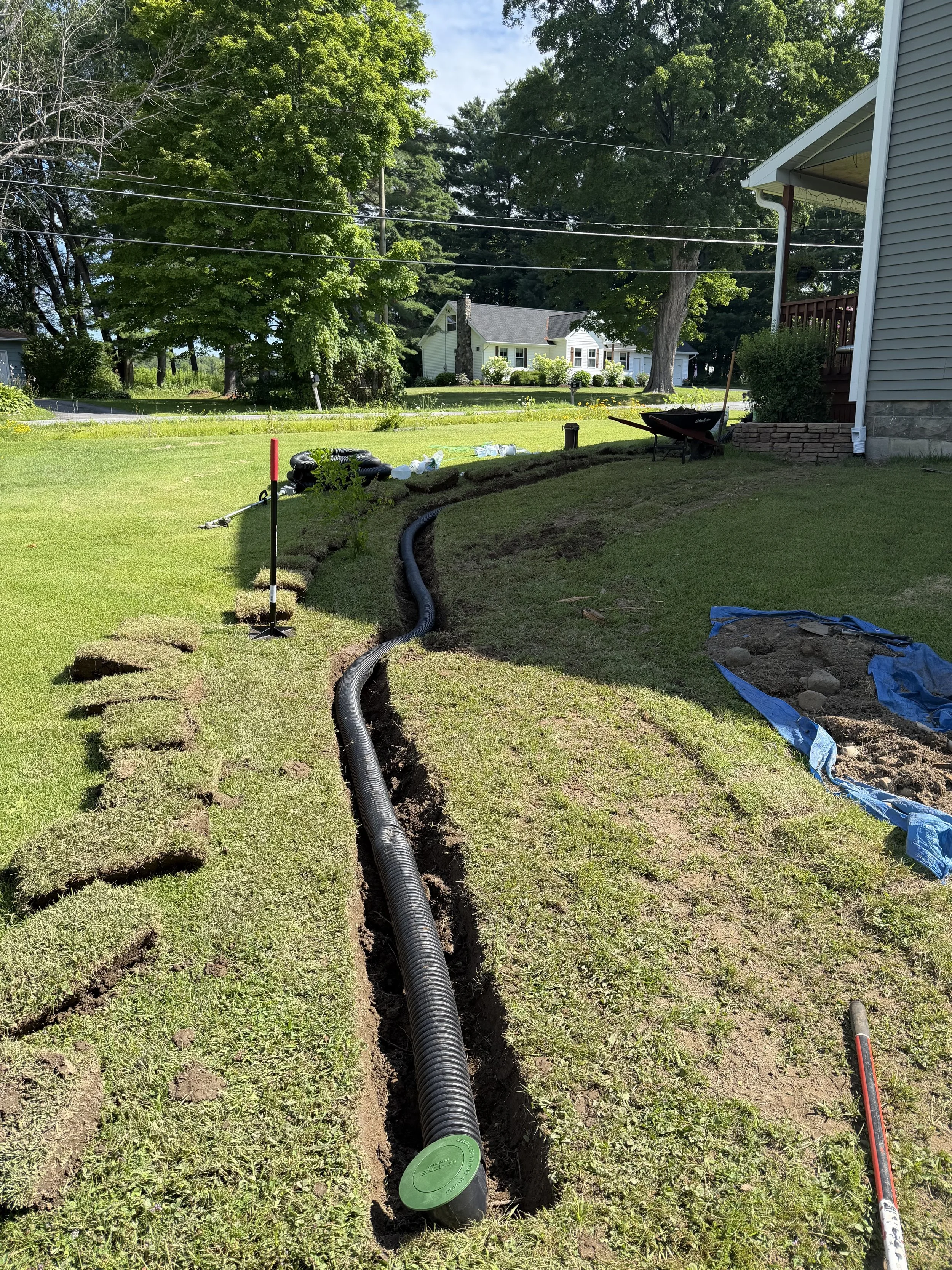 Flexible black conduit pipe installed underground in a trench in a grassy yard, with construction tools and supplies nearby, outside of a house with trees and neighboring homes in the background.