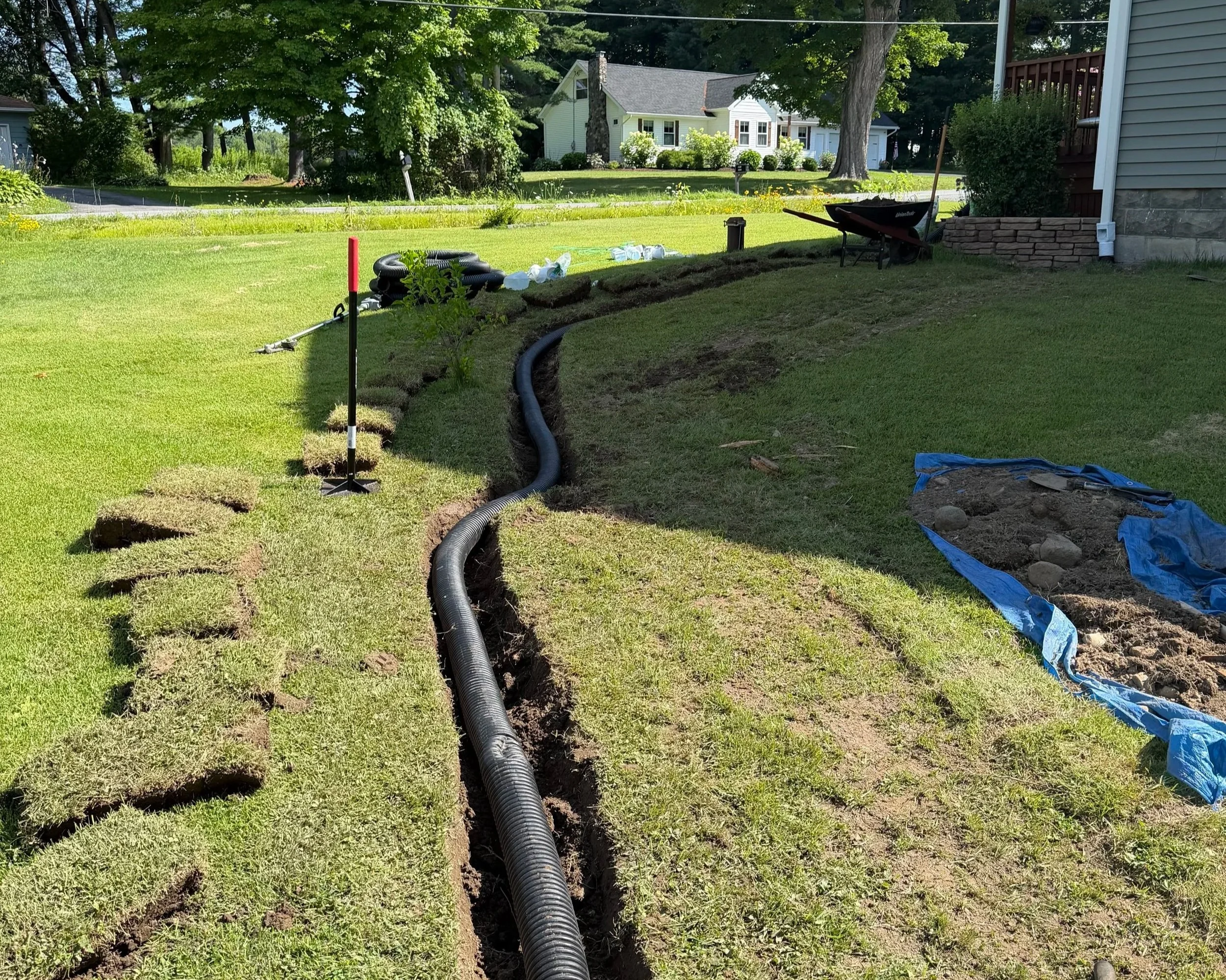 Lanscape view of a yard with trenching, black drainage pipes, and construction materials for yard drainage installation, with houses and trees in the background