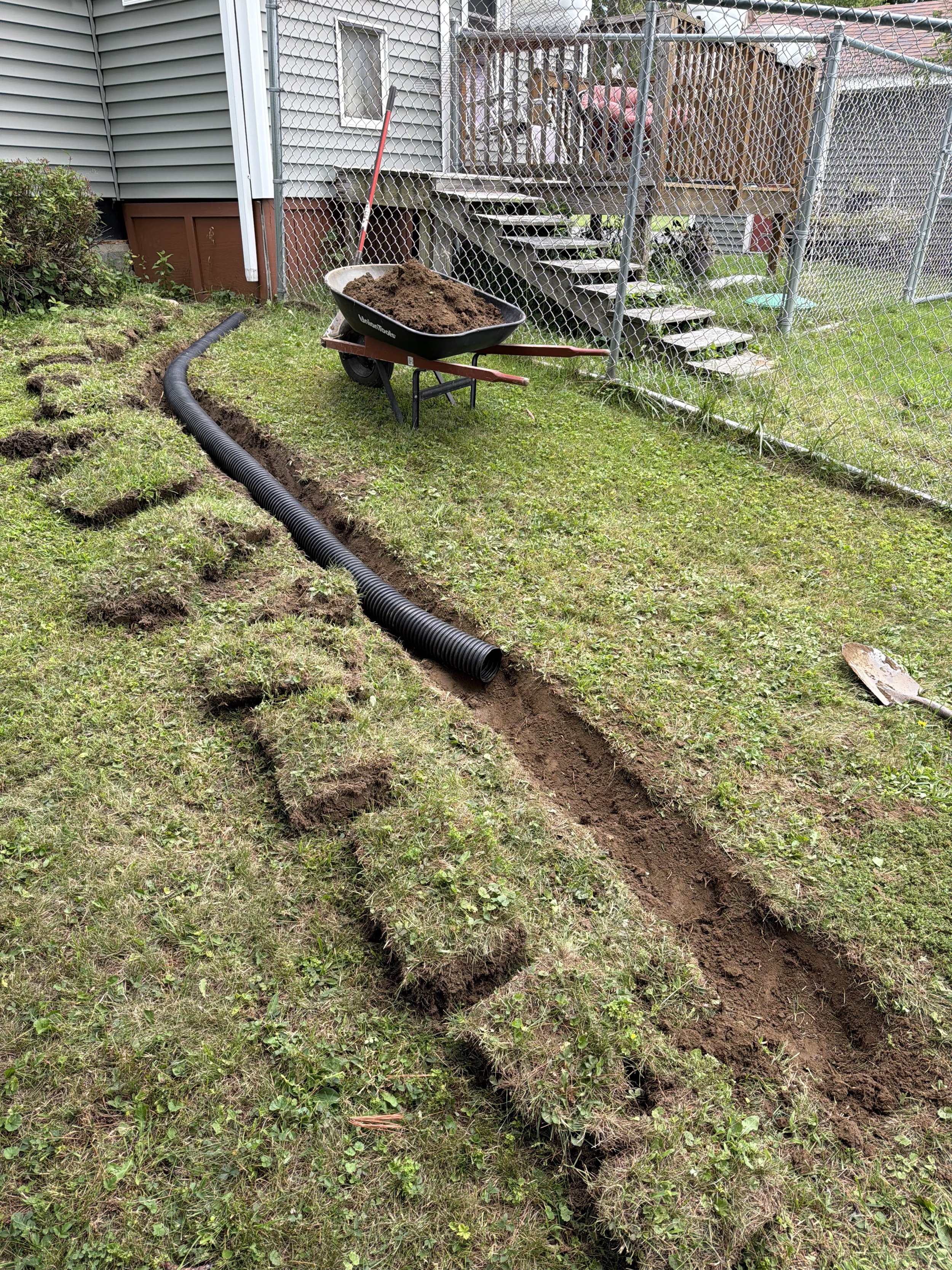 A trench dug in a grassy backyard with a black flexible drainage pipe placed along one side of the trench. A wheelbarrow filled with dirt is positioned near the trench, and a shovel is on the ground beside it. There is a chain-link fence and a raised wooden deck with stairs leading up to it in the background.