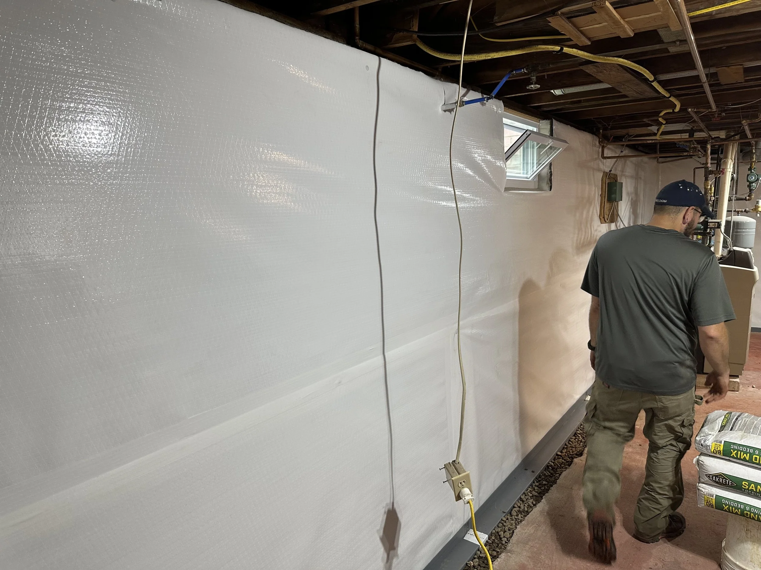 Two men working on a construction project inside a room with exposed ceiling beams and pipes. One man is wearing a cap and glasses, and the other is wearing a hat. There is a large white panel or wall, and various tools and materials around them.