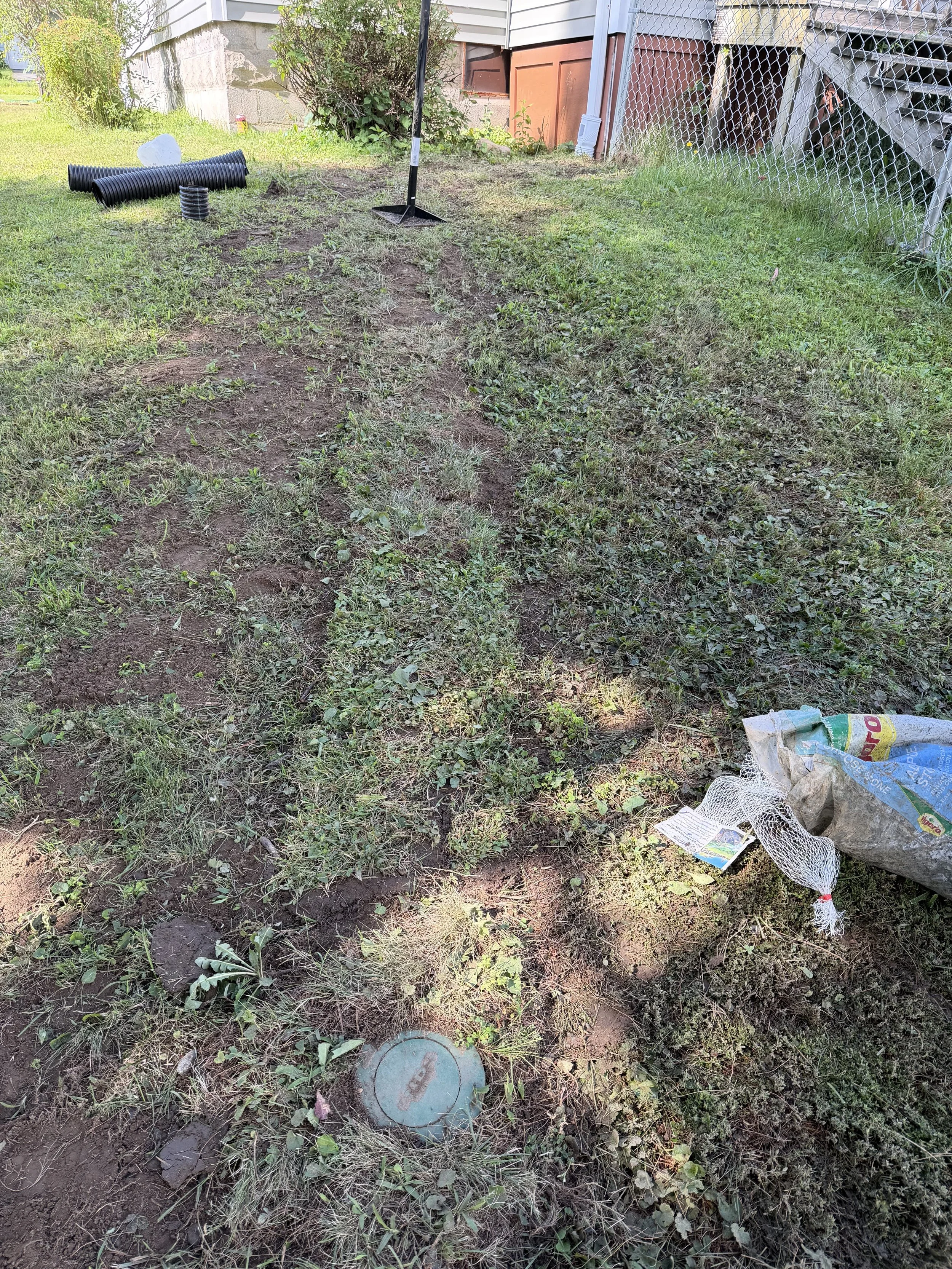A backyard with a patch of disturbed soil, a few small plants, and gardening tools including a rake. There are black corrugated pipes, a white plastic bag, and a mesh bag with damaged garden fencing nearby. The background shows parts of a house with siding, a chain-link fence, and some bushes.