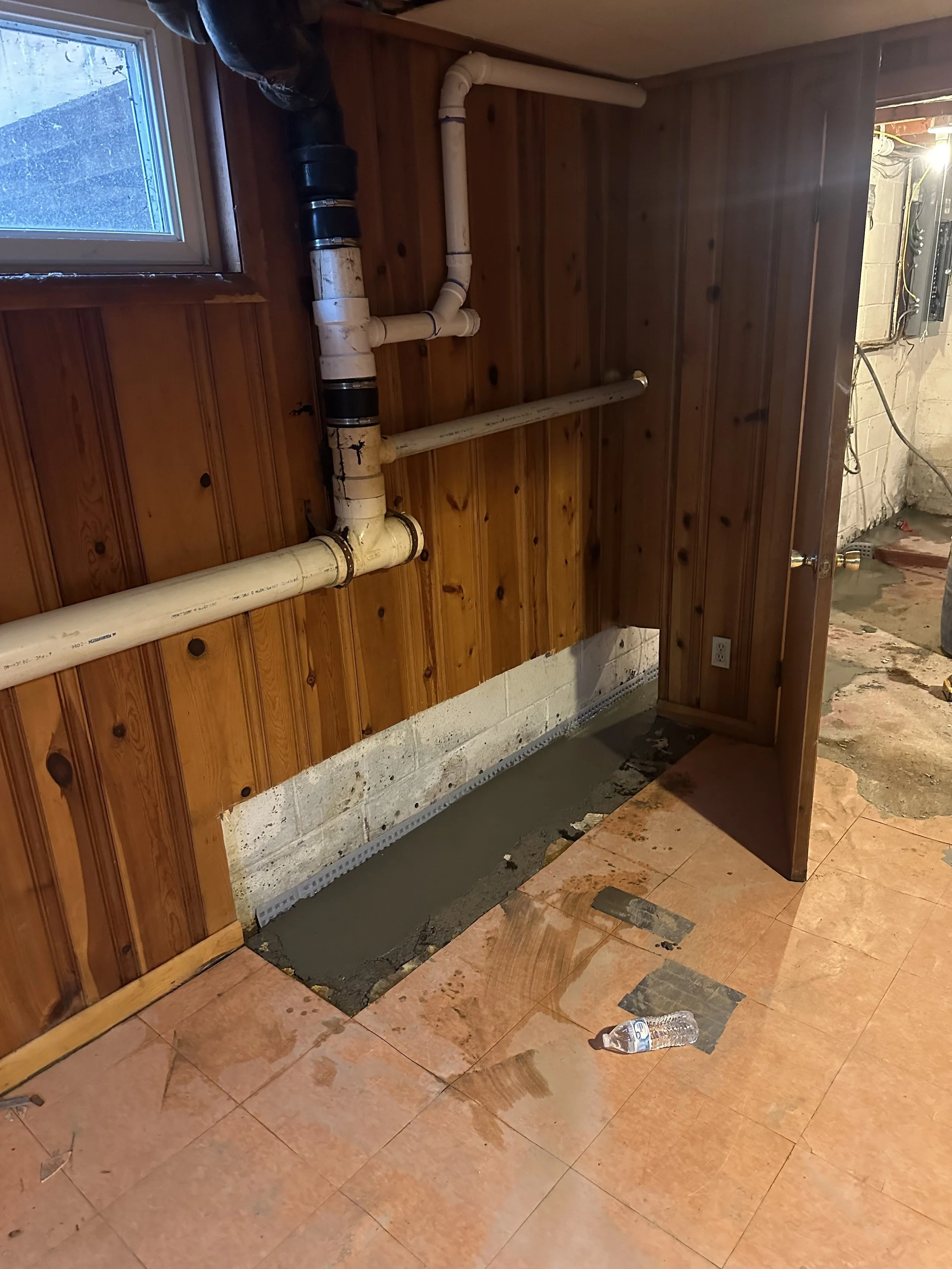 Construction area with exposed piping on a wood-paneled wall, a small window, and a partially tiled floor, with construction materials and a water bottle on the floor.