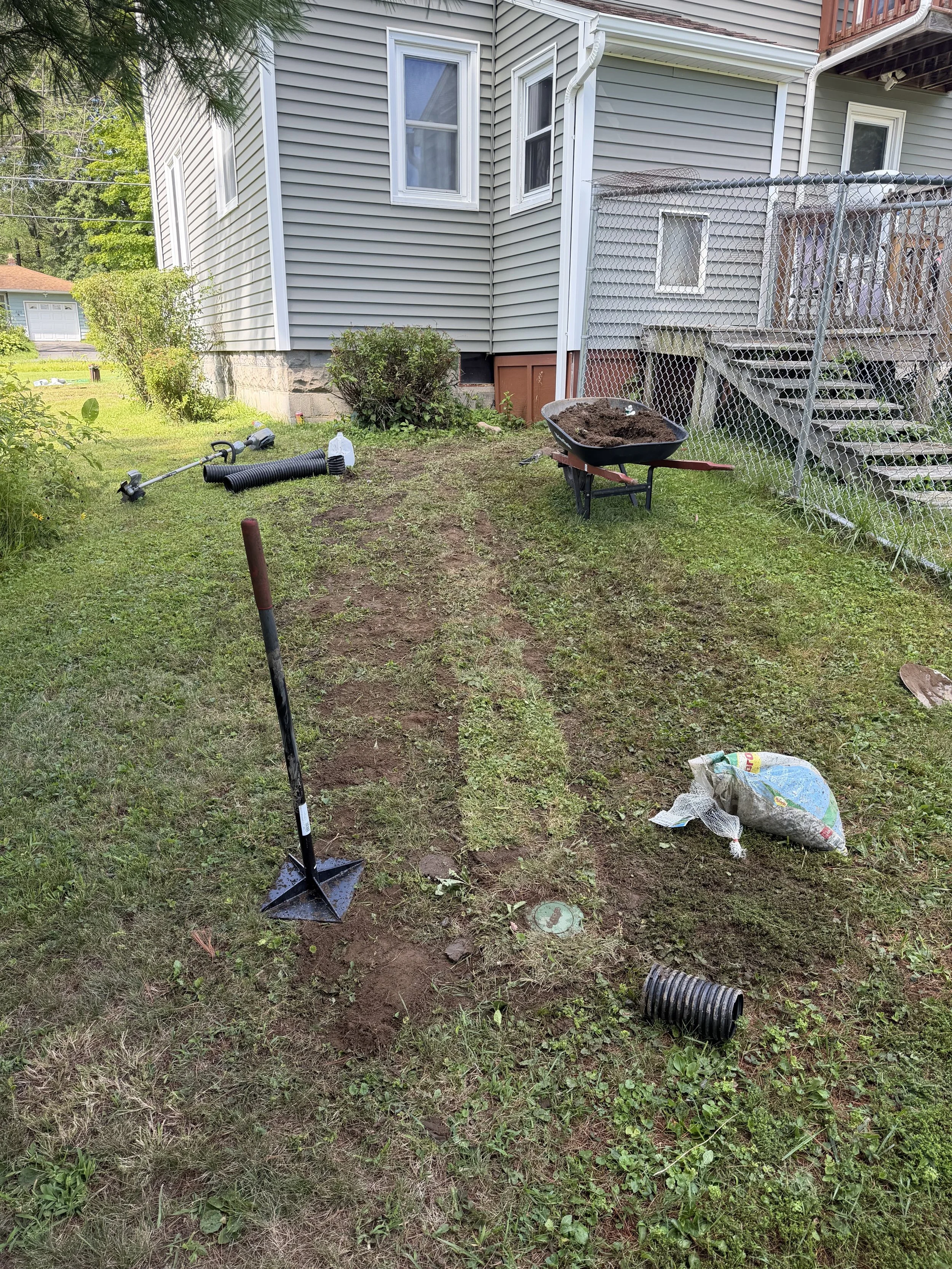 Backyard garden with a wheelbarrow filled with dirt, assorted gardening tools, a black drainage pipe, and patches of disturbed earth, with a house, fenced deck, and bushes in the background.