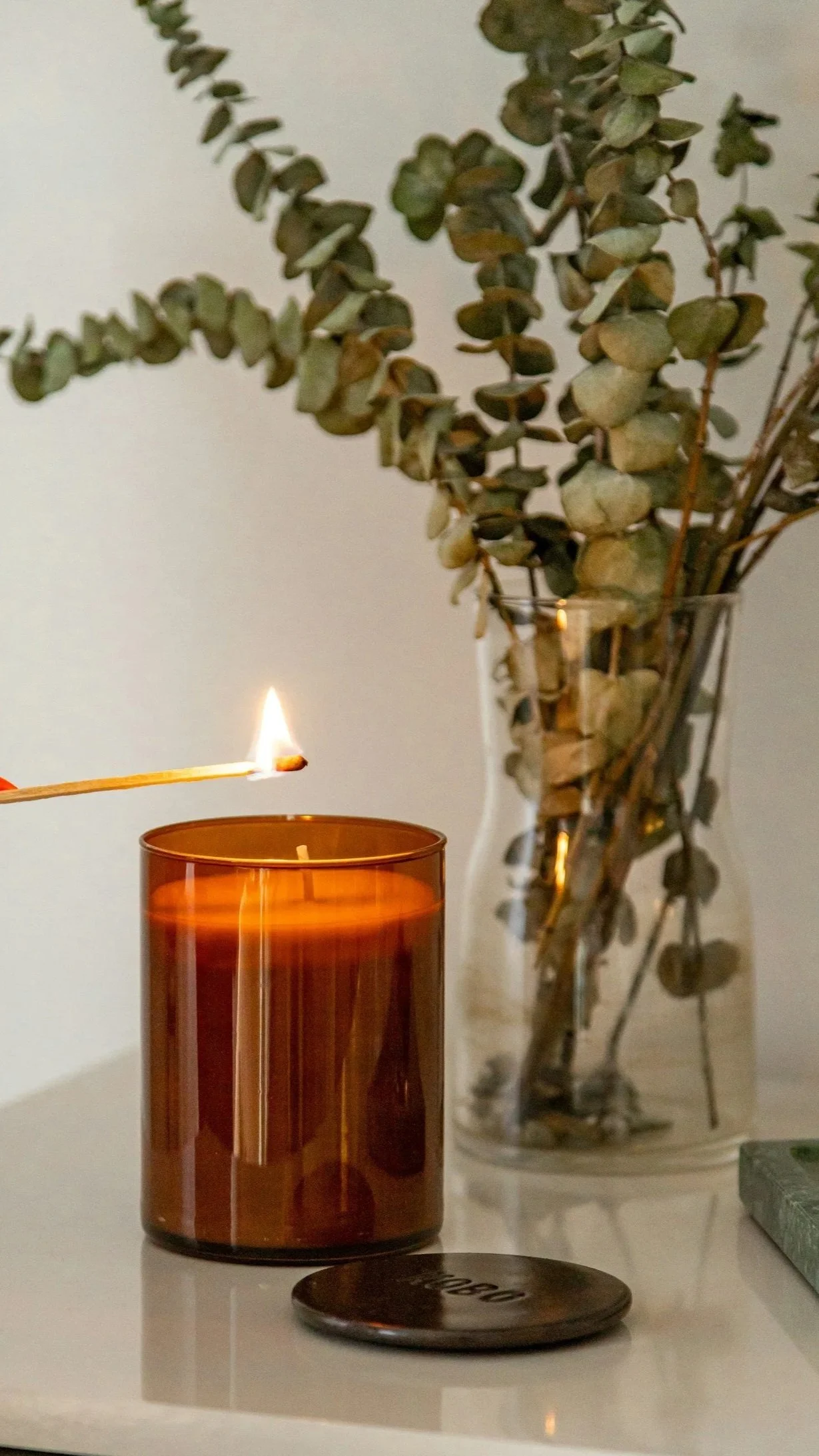 Lit match beside an amber glass candle and dried eucalyptus stems in a vase