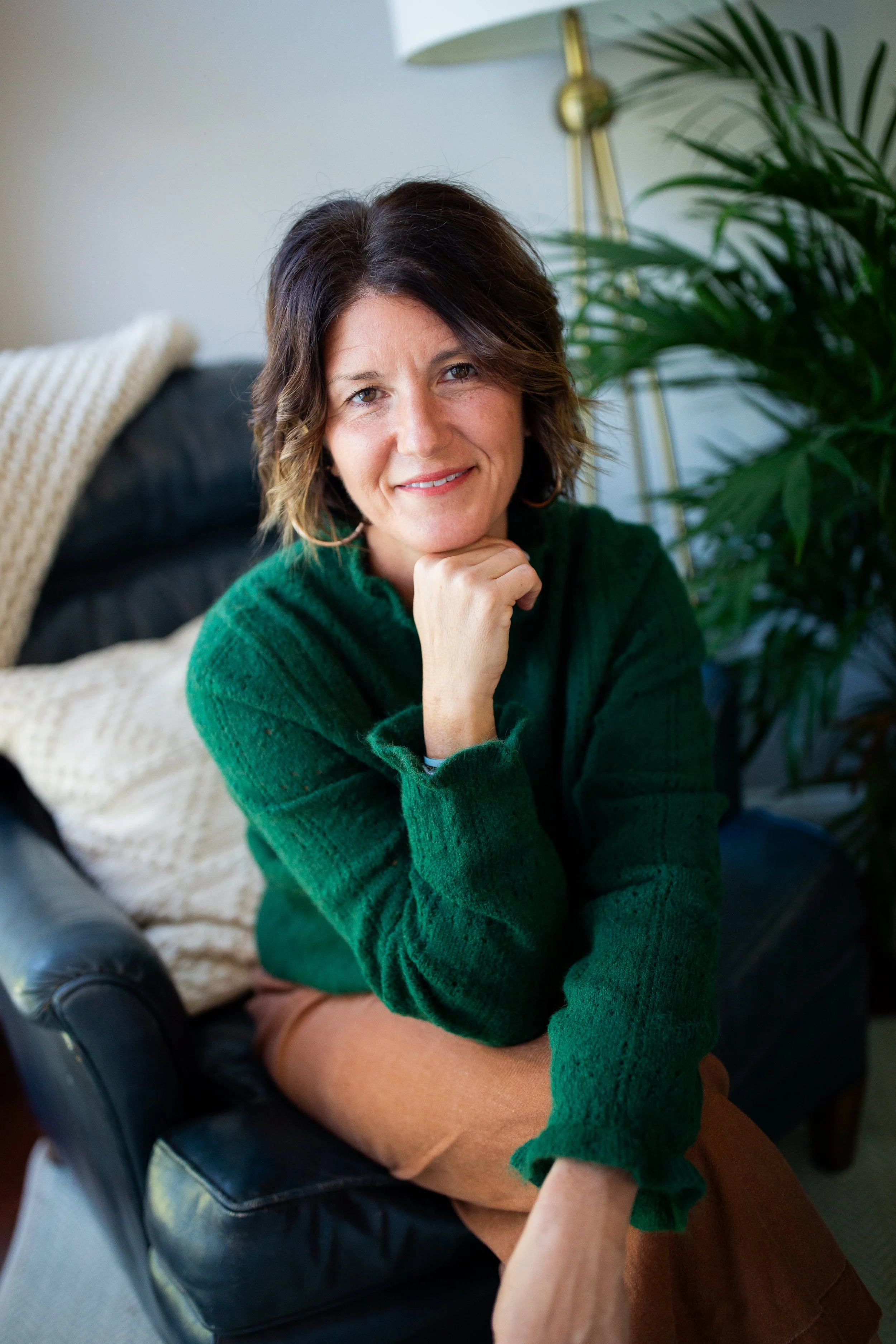 Smiling woman in a green sweater sitting indoors beside a large houseplant.