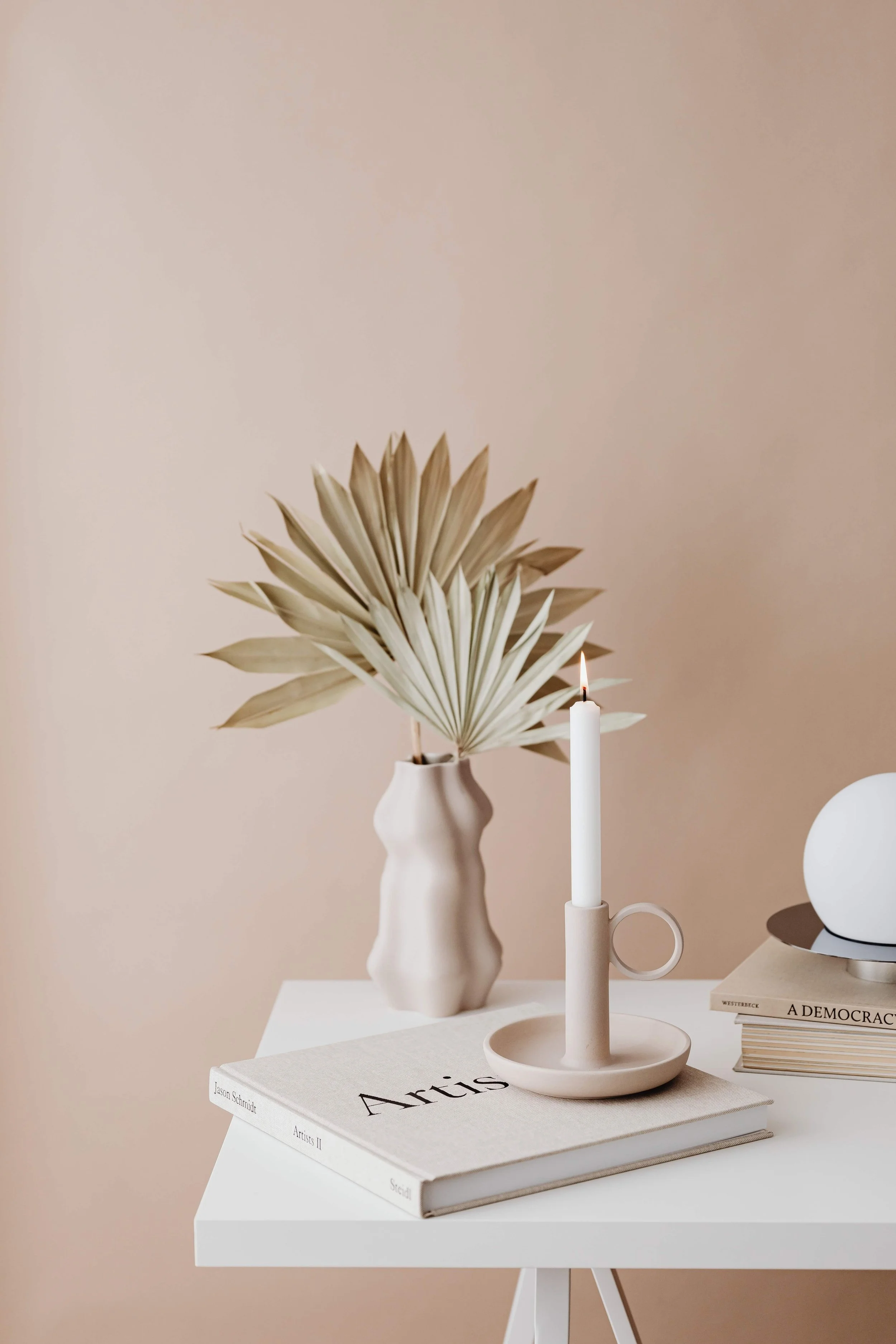 Minimalist home decor arrangement with dried palm leaves in a white vase, lit white candle, and stacked art books on a white table