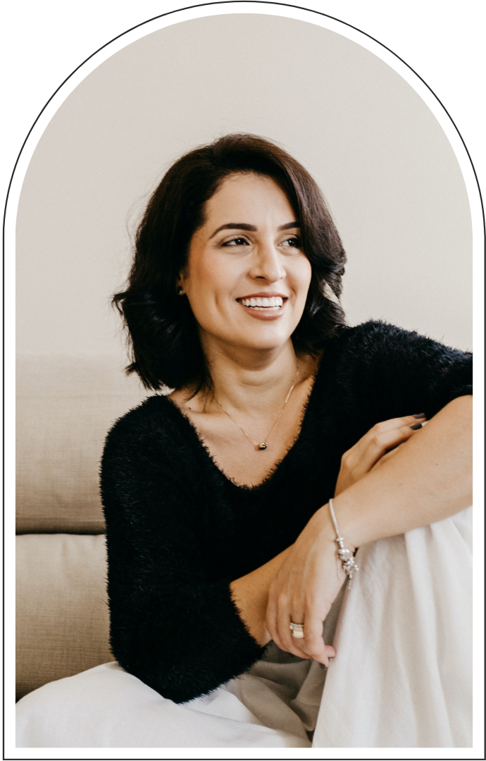 Smiling woman with dark hair wearing a black sweater and white pants, seated on a couch