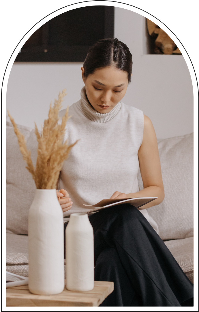 Woman in a white sleeveless turtleneck reading a book on a sofa beside a vase of dried flowers