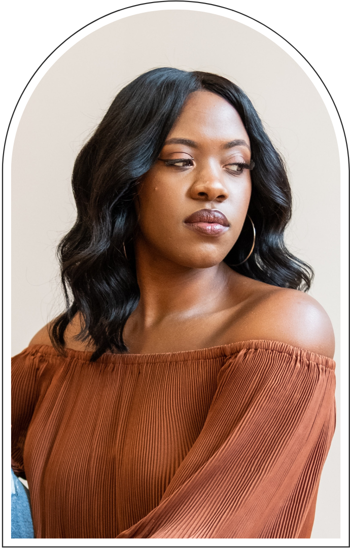 Portrait of a Black woman with wavy black hair and hoop earrings wearing a rust-colored off-the-shoulder pleated blouse
