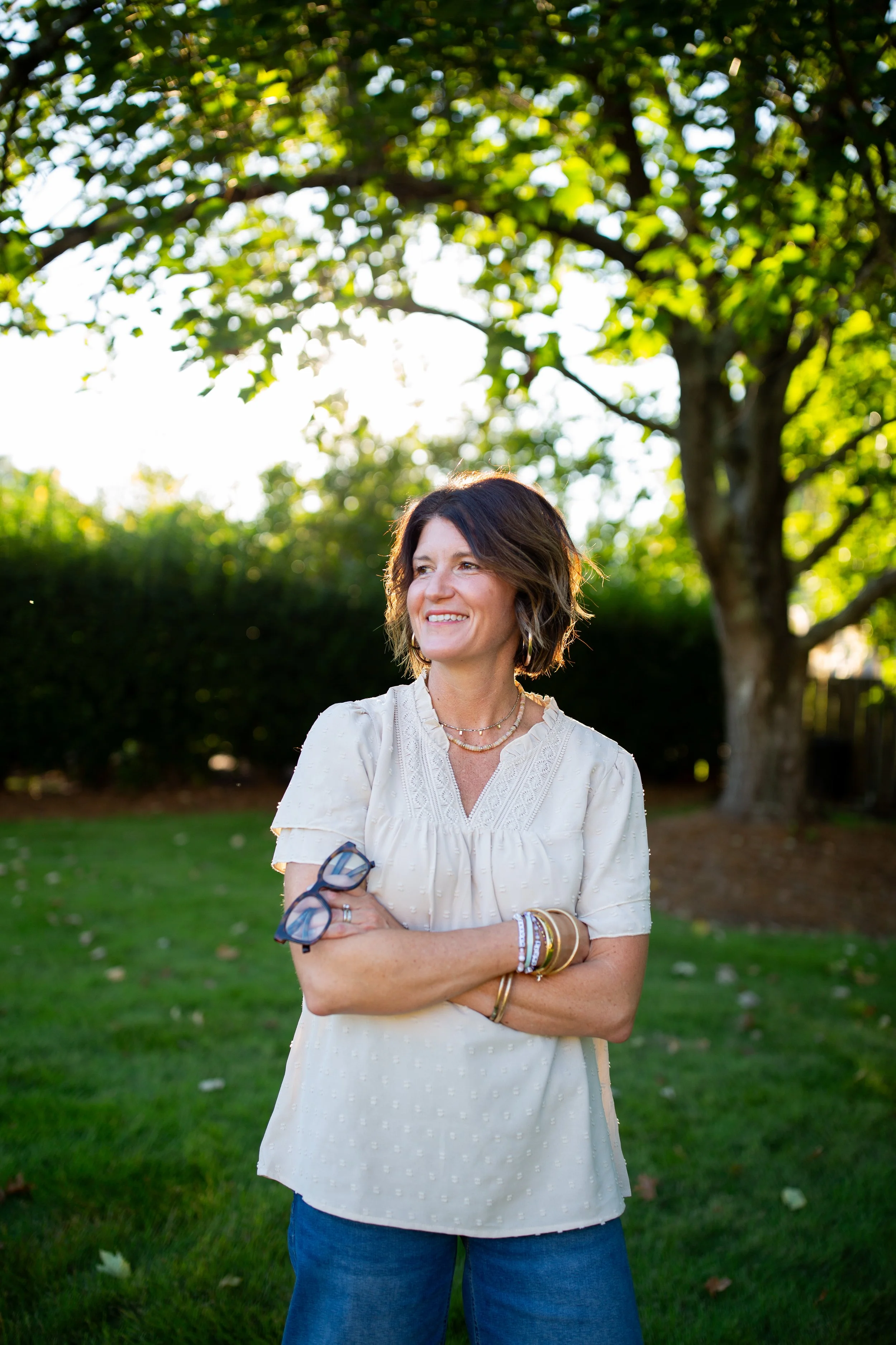 Smiling woman in a white blouse standing outdoors in a sunlit garden, holding eyeglasses.