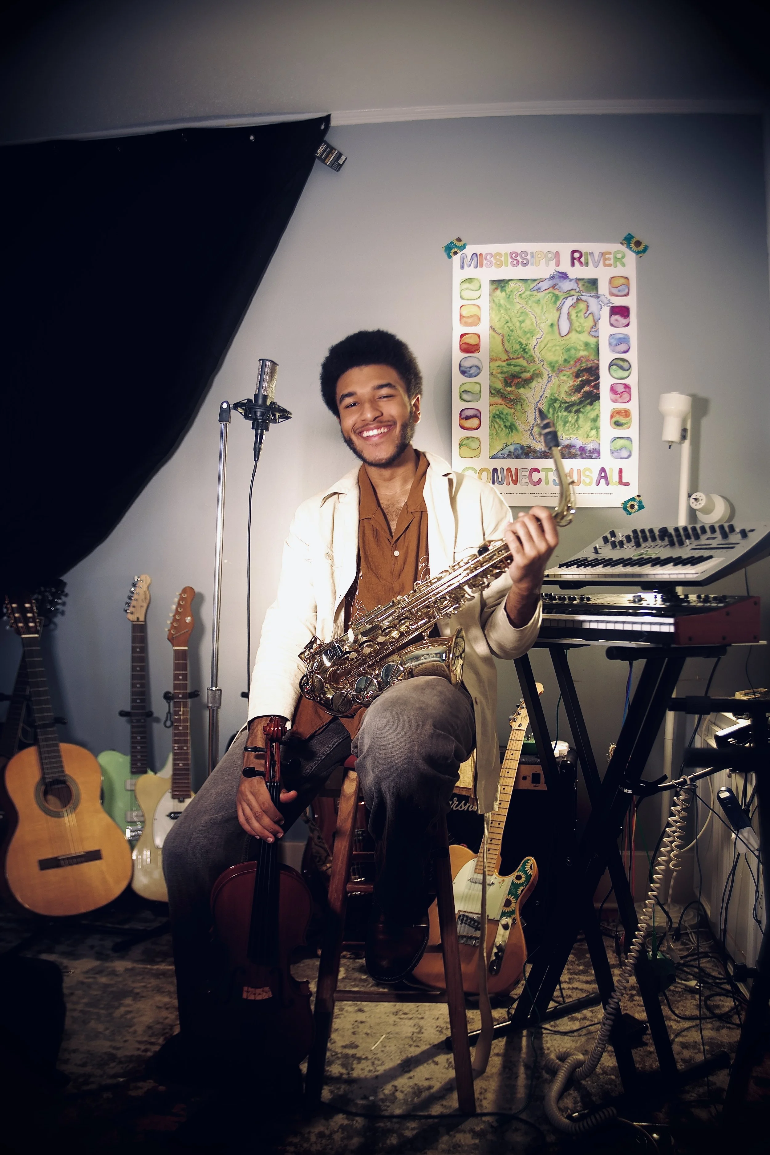 A young man with an afro hairstyle and a beard, smiling, holding a saxophone, sitting in a music studio surrounded by guitars and musical equipment, with a poster of the Mississippi River on the wall.