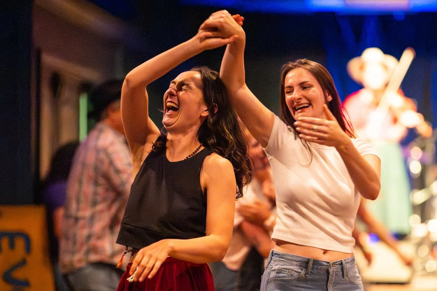 Two women are dancing happily at a party or celebration, with one woman holding her hand high and the other smiling. Background shows other people and a colorful, festive setting.
