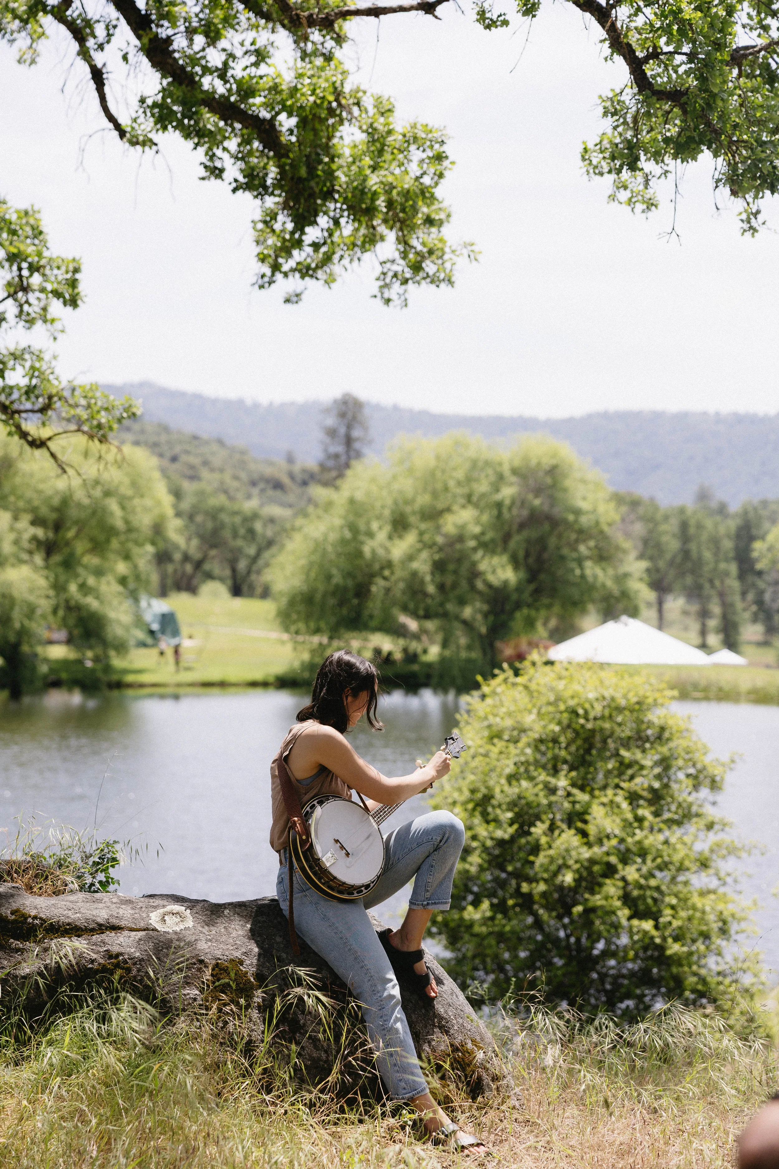 A young woman sits on a rock by a lakeside, playing a banjo, with trees and hills in the background on a sunny day.