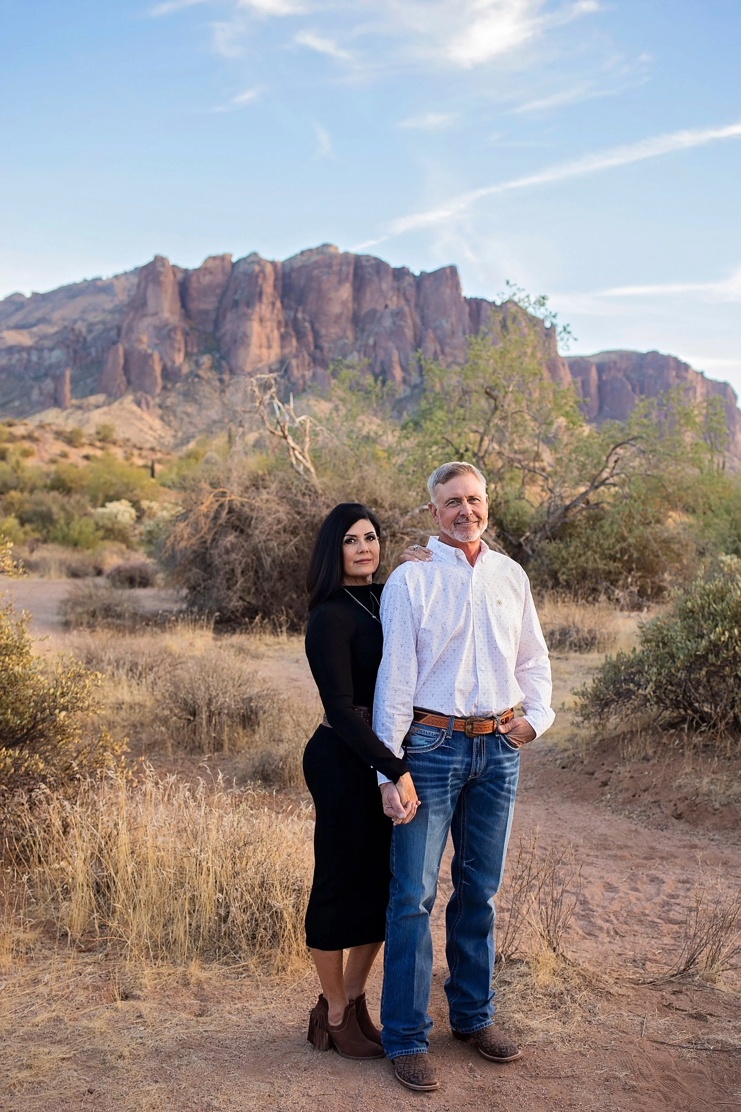A couple standing in a desert landscape with mountains and sparse vegetation in the background. The woman has dark hair and is wearing a black dress with brown ankle boots. The man has gray hair and a beard, wearing a white patterned shirt and blue jeans.
