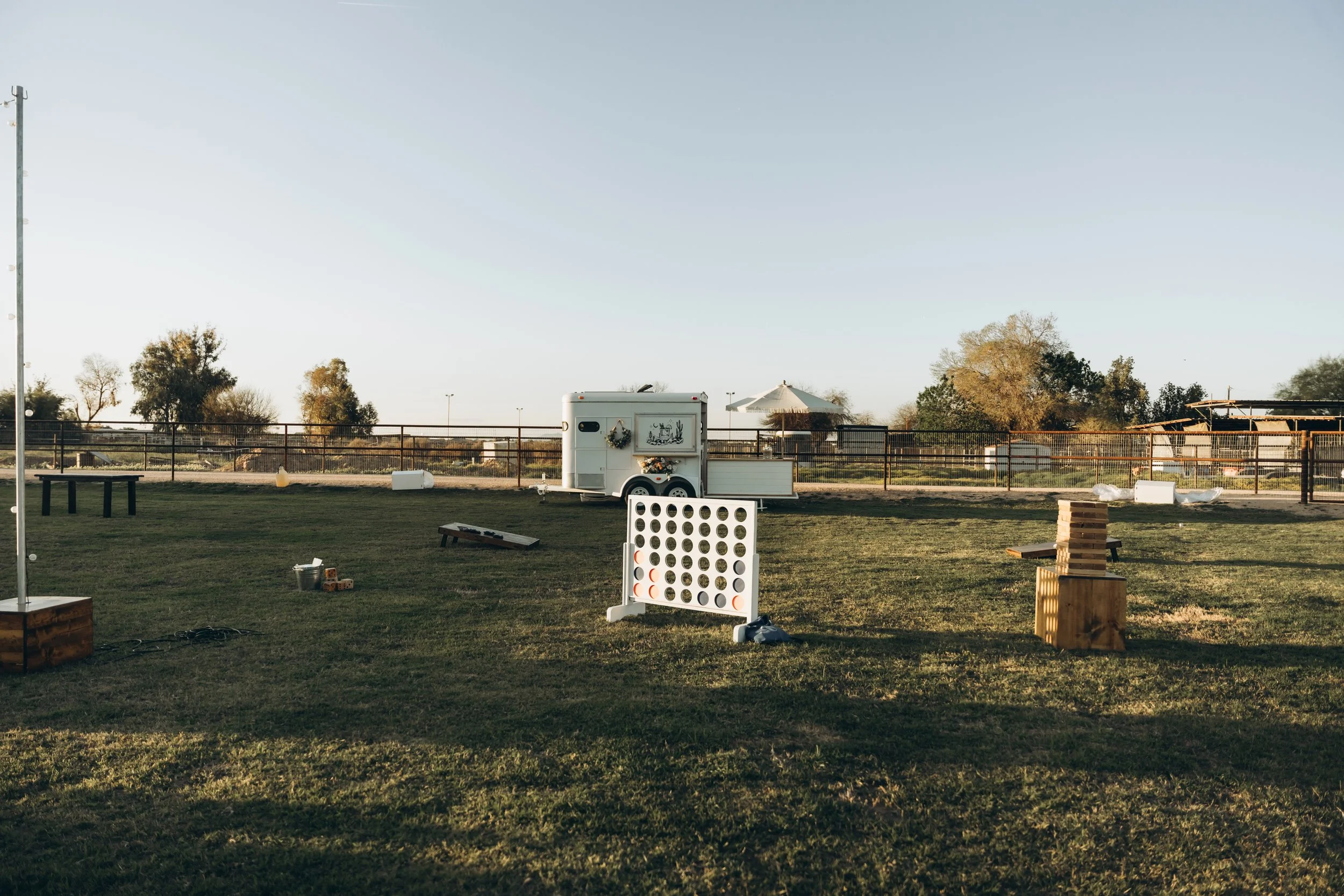 An outdoor recreational area with a grassy field, a large outdoor connect four game, various wooden blocks, plastic buckets, and a popcorn trailer, surrounded by a fence and some trees under a clear sky.