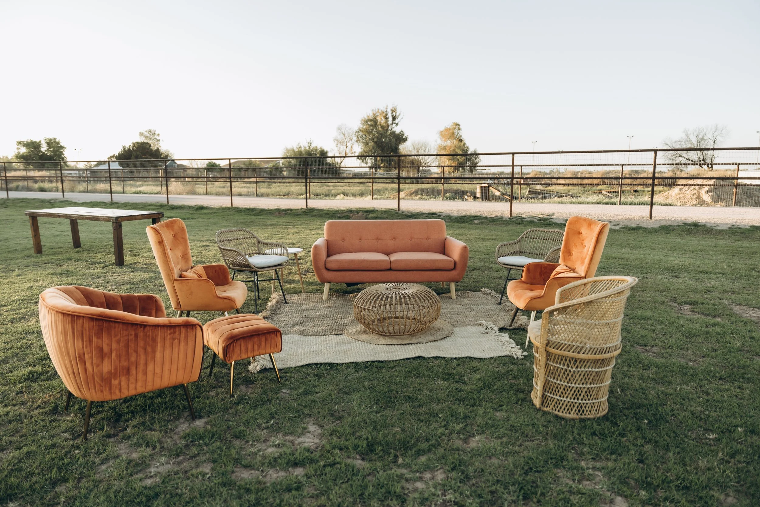 Outdoor sitting area with peach-colored sofa, velvet armchairs, wicker chairs, and a round wicker coffee table on a rug on grass, with a fence and trees in the background.