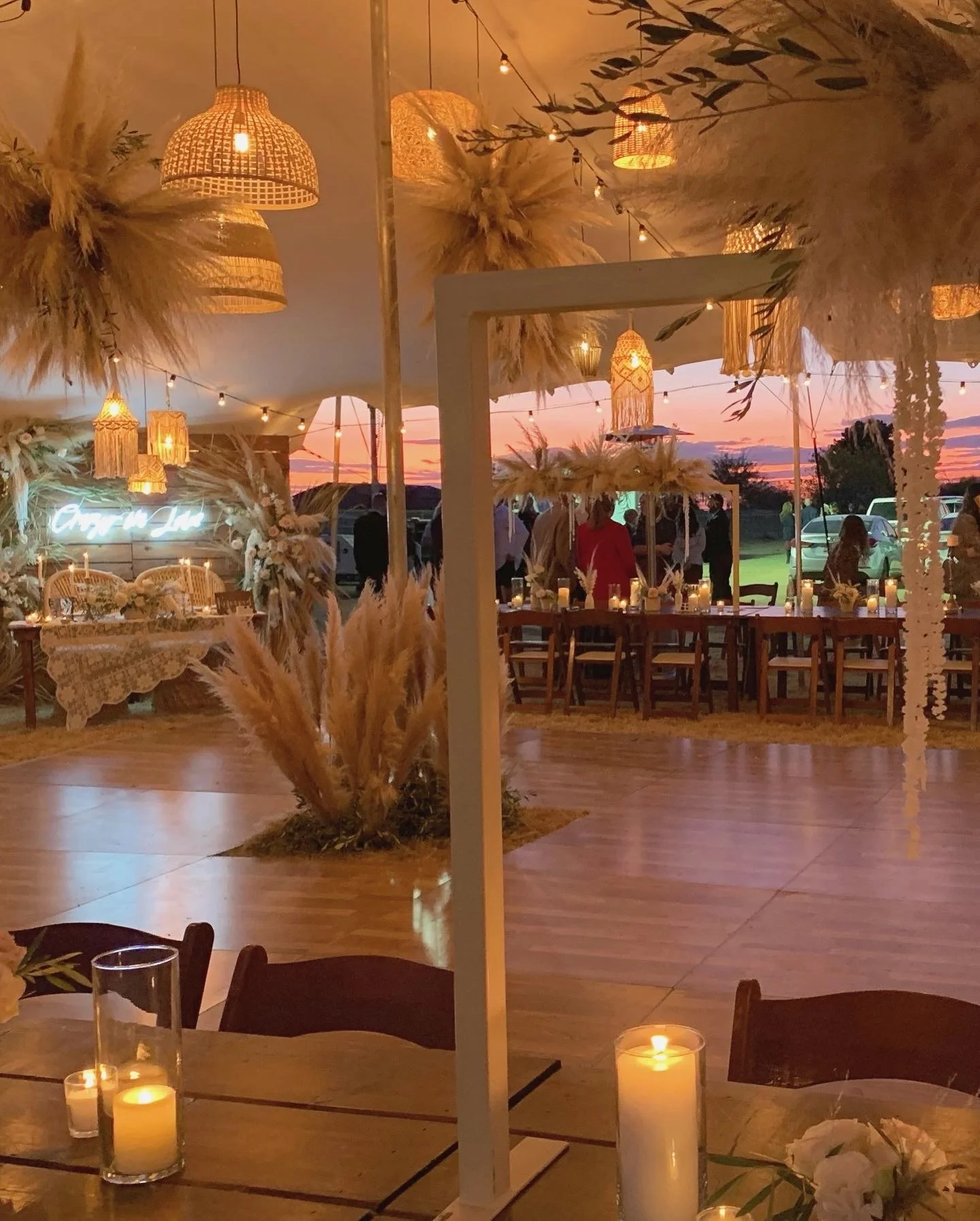 An outdoor evening event space decorated with warm string lights, lanterns, and candles. There are tables, chairs, and floral decorations, with a sunset sky in the background.