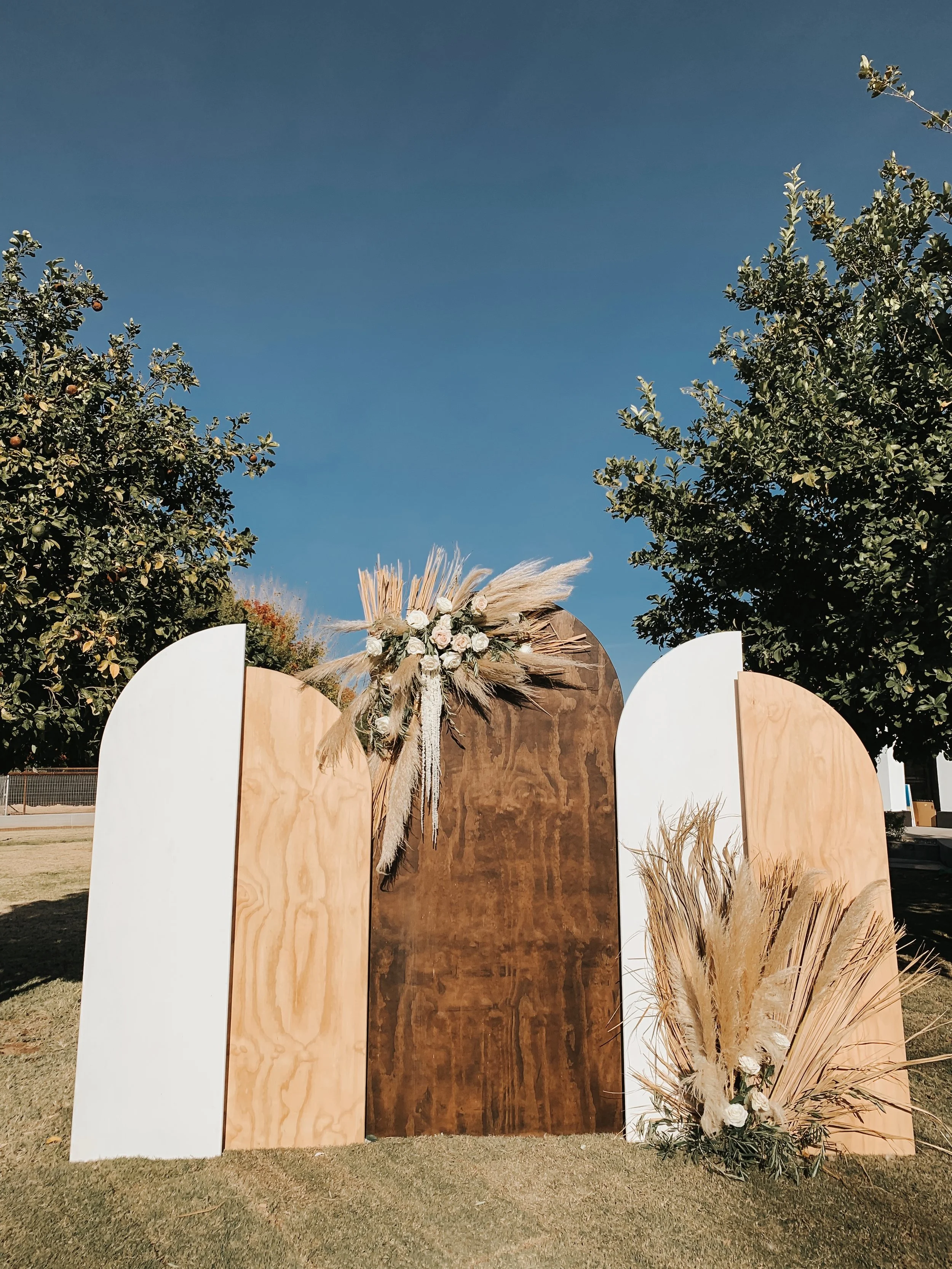 Decorative backdrop for an outdoor event featuring large white and brown panels with floral and pampas grass arrangements, set against a clear blue sky and green trees.