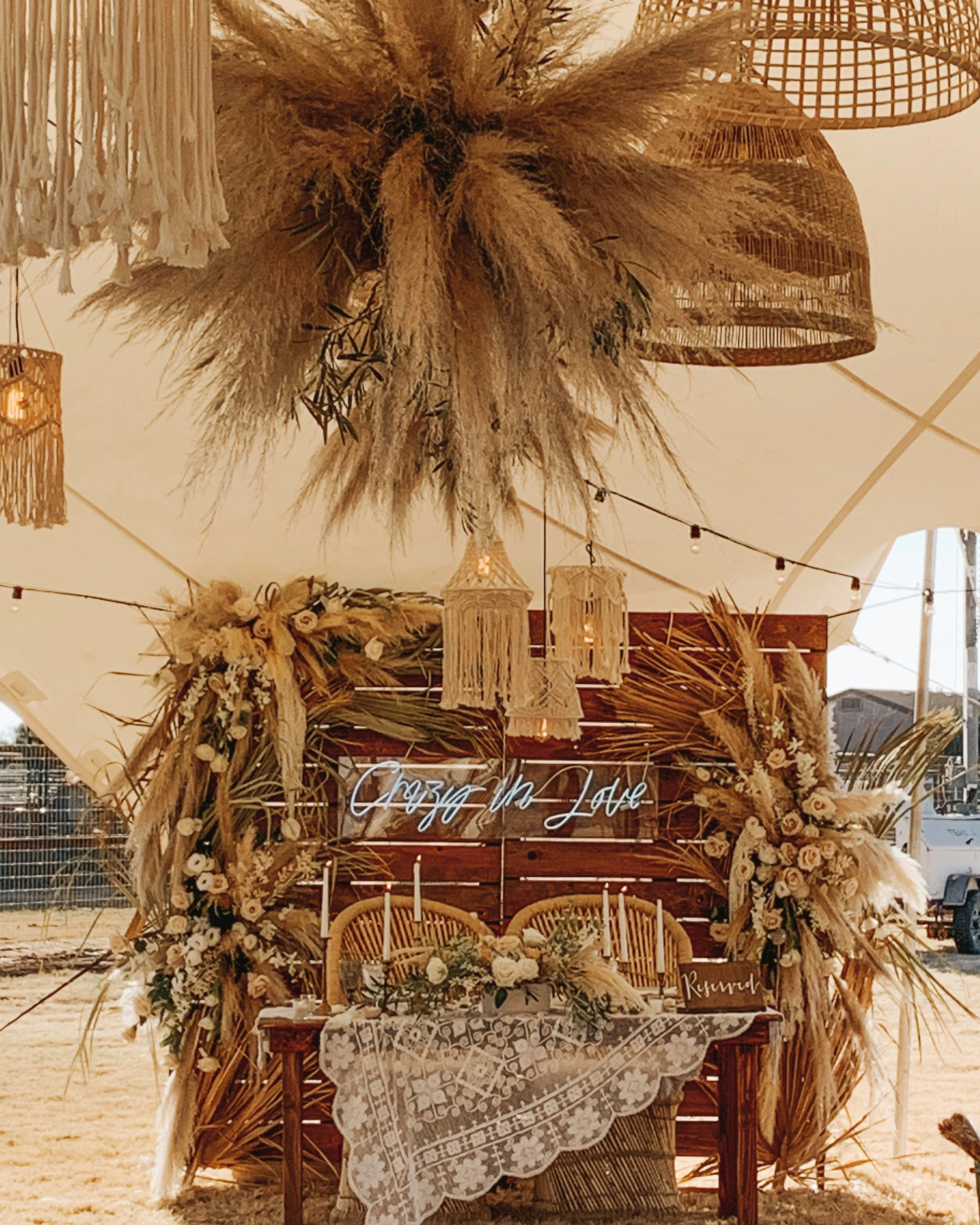 A wedding altar decorated with dried grass, flowers, and rattan elements. A wooden table with a lace cloth is in front, with candles and floral arrangements. White neon sign says 'Crazy In Love'.