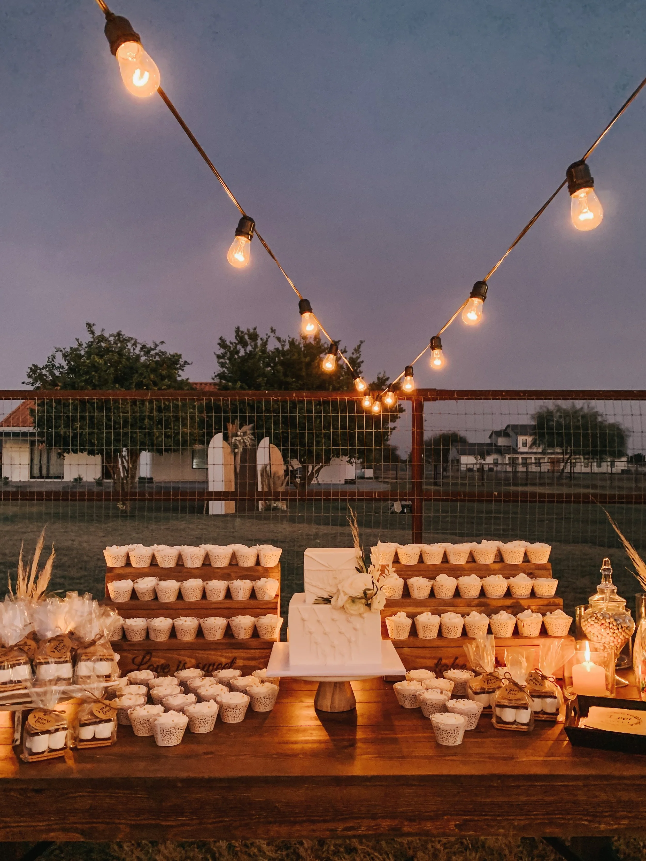 Outdoor evening dessert table with a rustic wooden setup, decorated with cupcakes arranged on wooden stands, a white cake, and small jars; string lights hanging overhead create a warm ambiance.